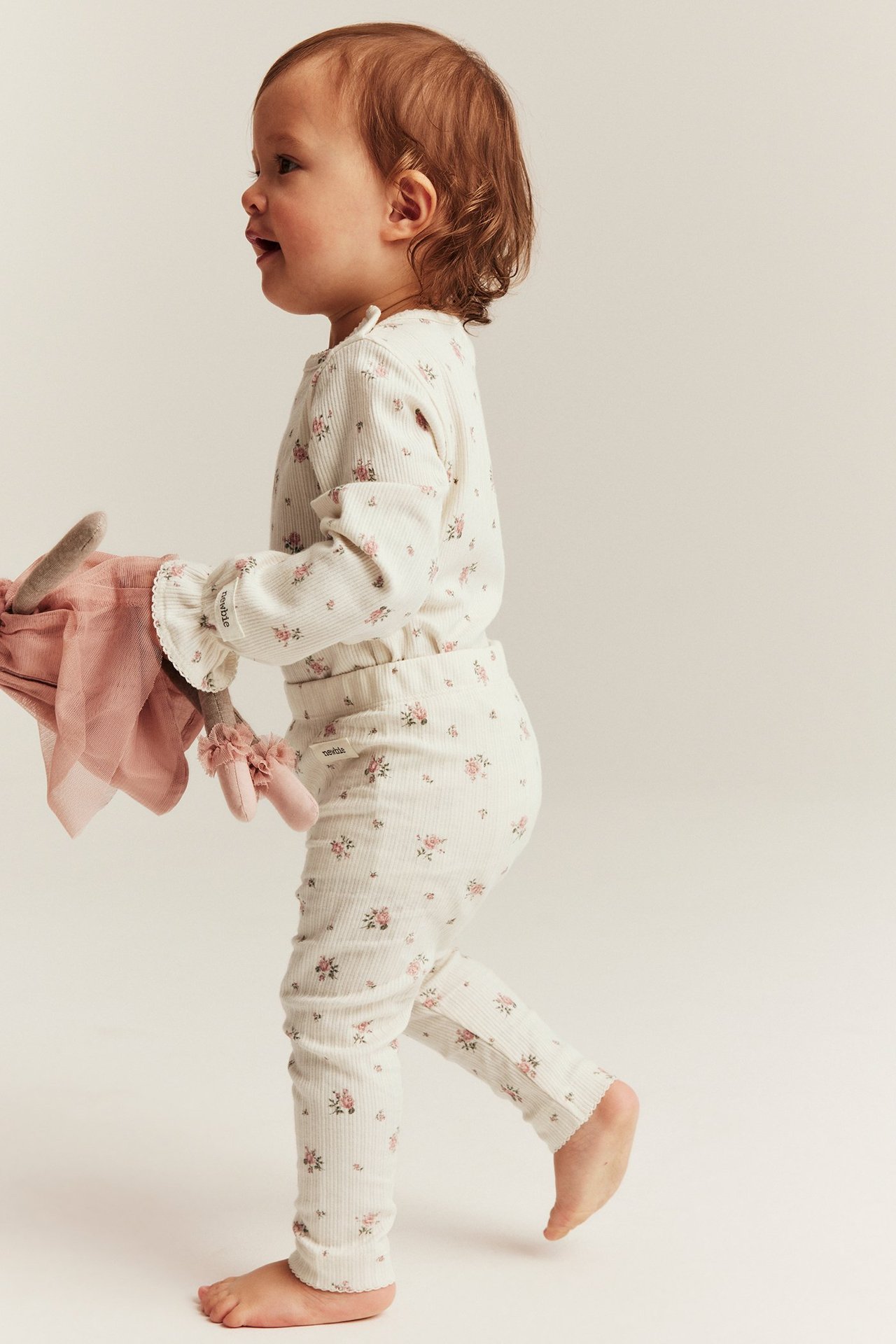 Widok z boku dziecka w kremowych prążkowanych legginsach w kwiaty i pasującym topie, trzymającego różową lalkę.