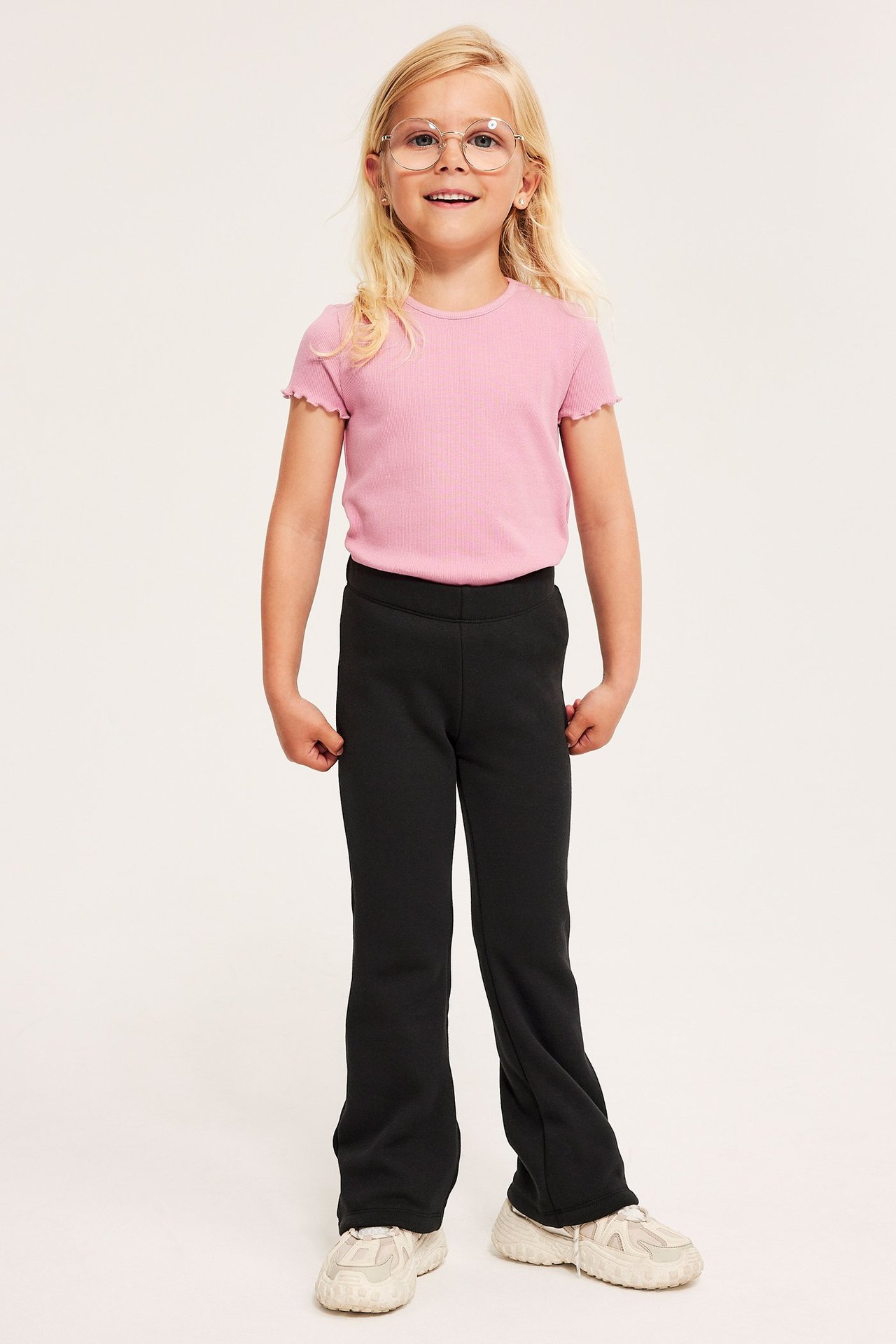 Edestä: lapsi mustissa levenevissä leggingseissä, vaaleanpunaisessa ribbipaidassa, pyöreissä laseissa, valkoisissa lenkkareissa.