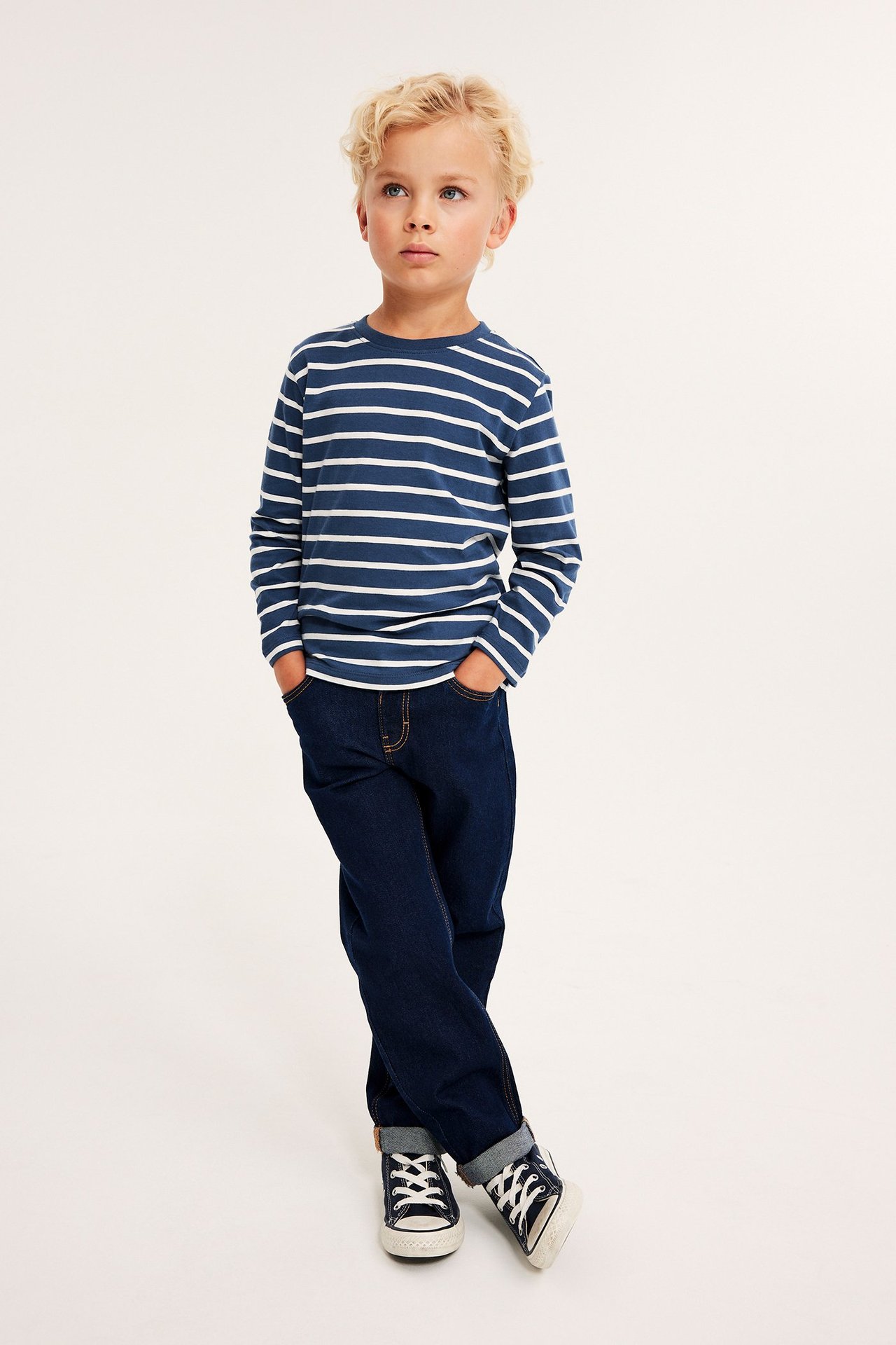 Edestäpäin, lapsi tummansinisessä ja valkoisessa raidallisessa pitkähihaisessa t-paidassa, tummansinisissä farkuissa ja lenkkareissa.