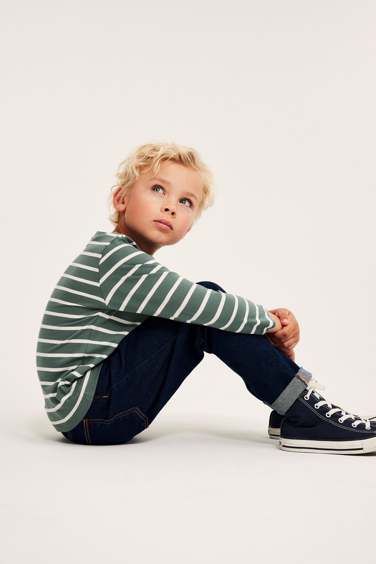 Widok z boku dziecka w ciemnozielono-białej koszulce z długim rękawem, ciemnoniebieskich dżinsach i trampkach.