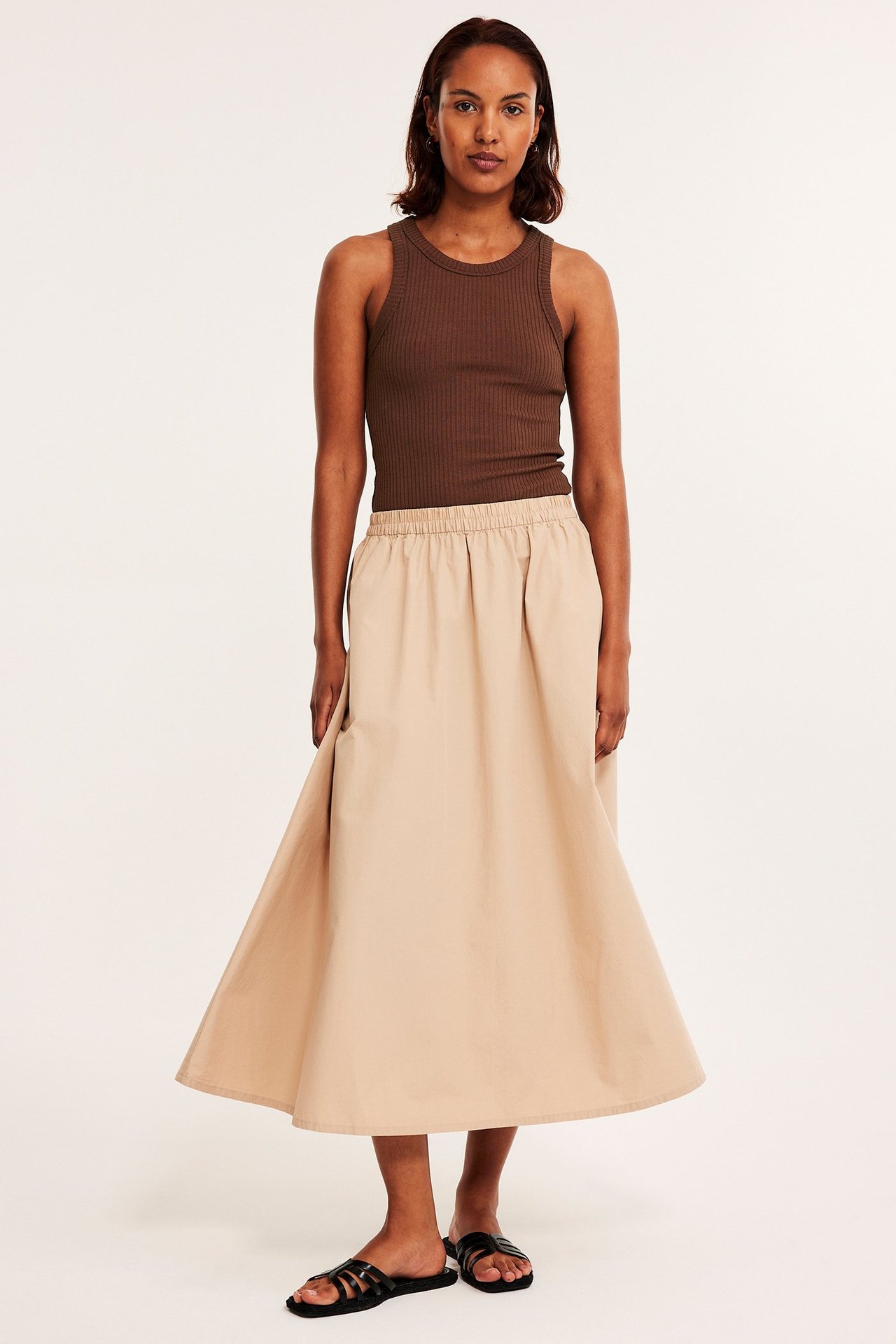 Forfra: kvinne i brun ribbet singlet, beige midiskjørt og svarte flate sandaler.