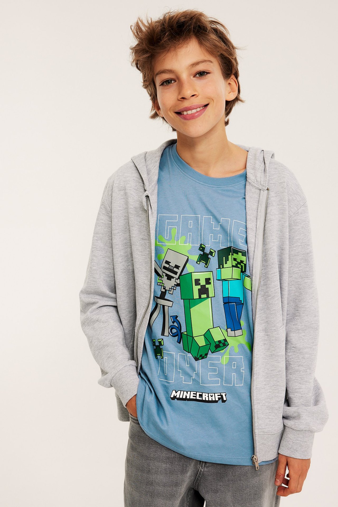 Framifrån: Barn bär blå bomulls-T-shirt med Minecraft-tryck, under öppen grå dragkedjeförsedd huvtröja.
