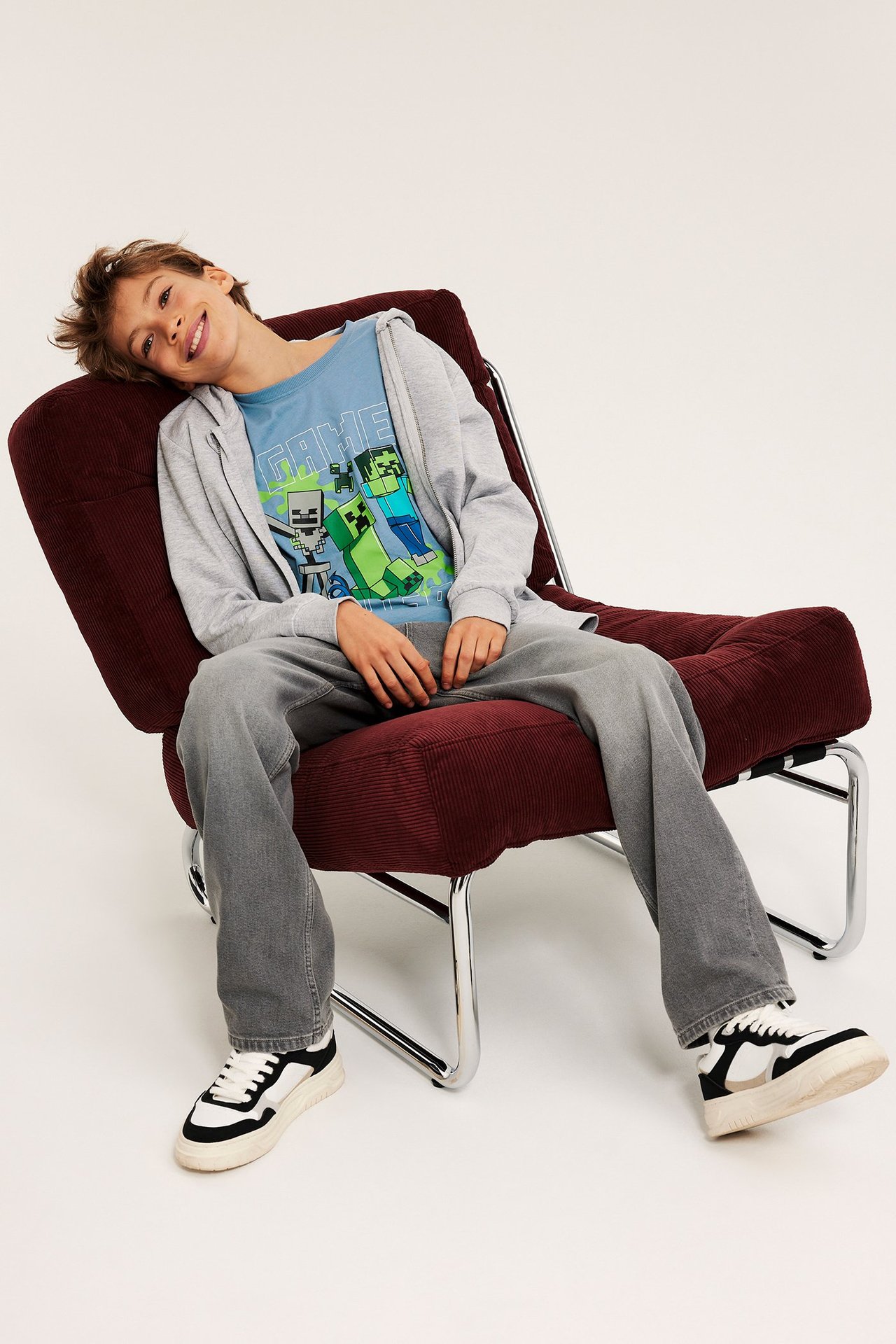 Barn i blå Minecraft-print T-skjorte, lys grå hettegenser med glidelås, grå bukser og svarte og hvite sneakers.