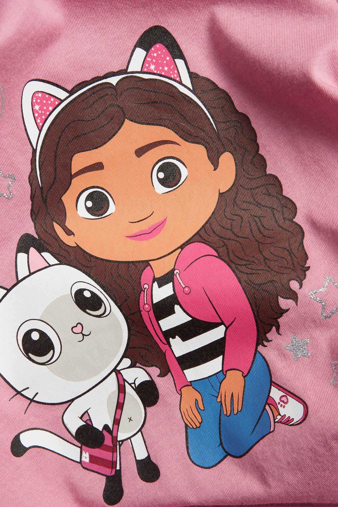 Zbliżenie różowej bawełnianej koszulki dziecięcej z długim rękawem, z nadrukiem Domku Gabi i brokatowymi gwiazdkami.