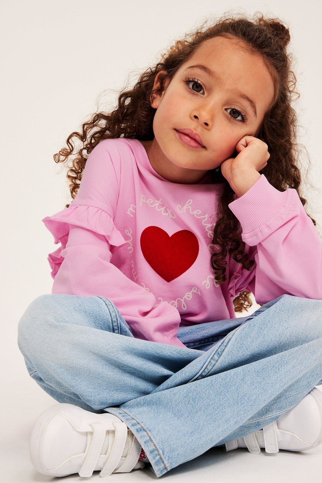 Widok z przodu: dziecko w jasnoróżowej bluzie z czerwonym sercem, falbankami, jasnoniebieskich dżinsach i białych trampkach.