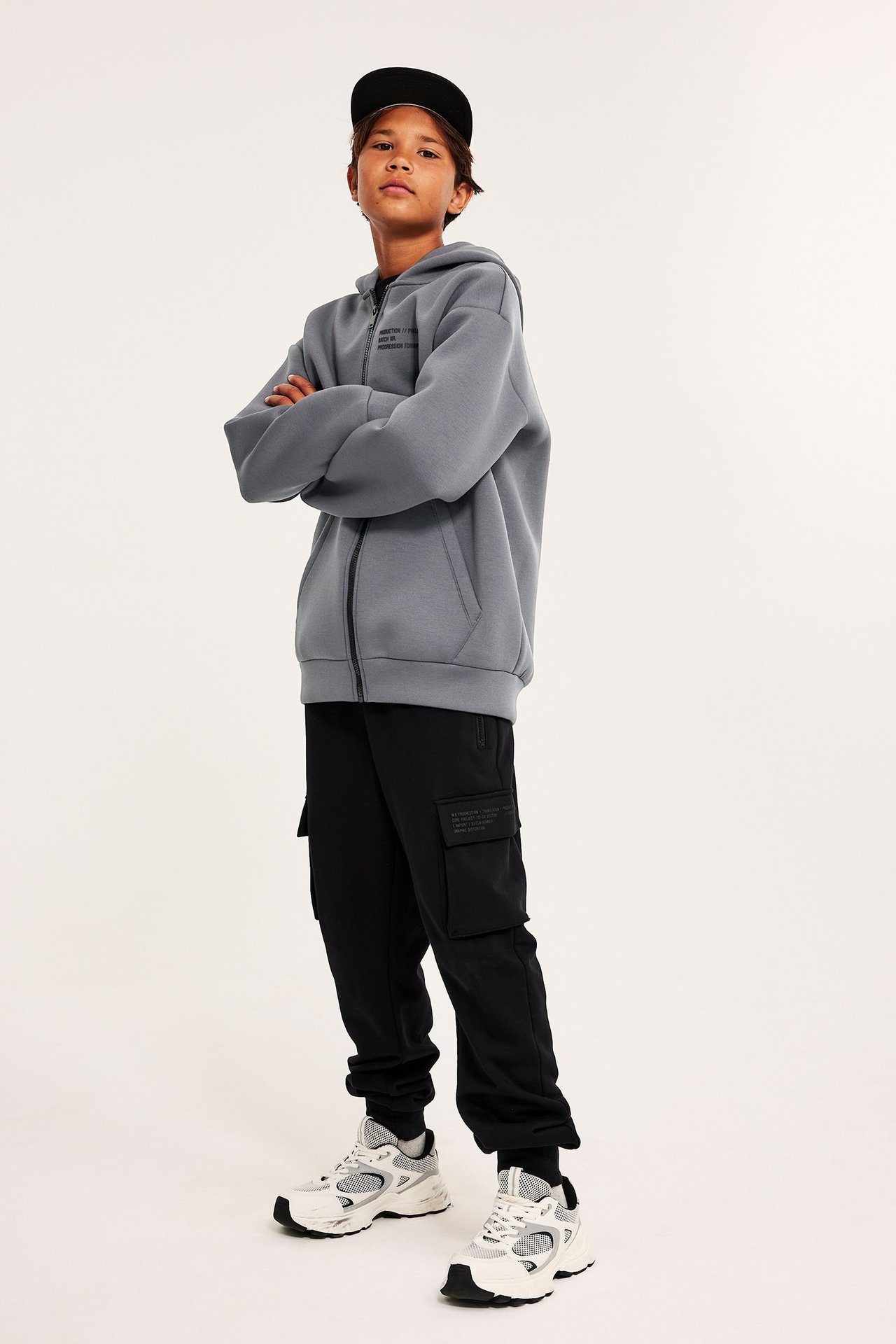 Widok z przodu: Dziecko w szarej bluzie scuba, czarnych spodniach cargo, białych sneakersach i czapce.