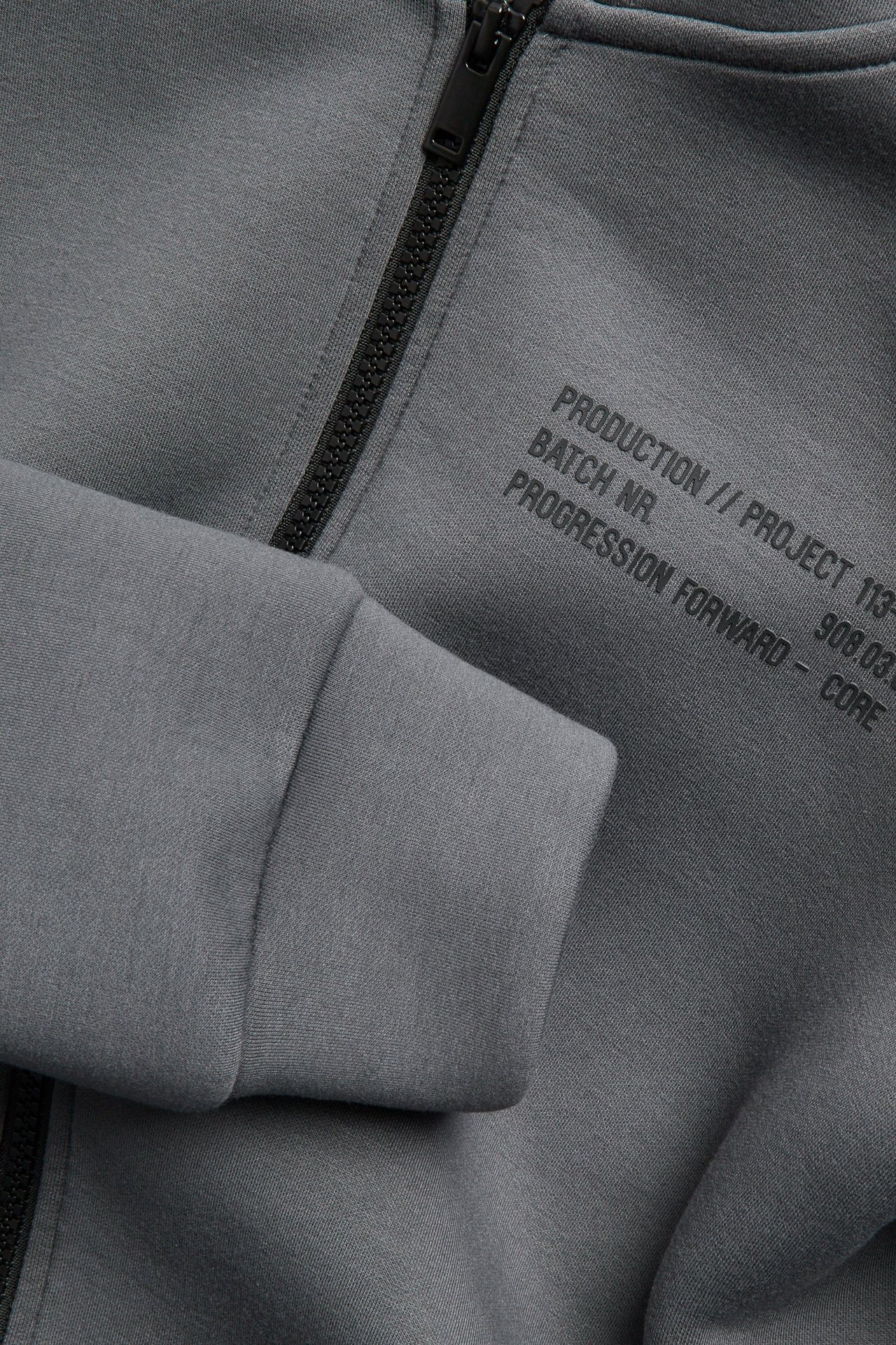 Zbliżenie szarej bluzy z kapturem z materiału scuba, z czarnym zamkiem i czarnym nadrukiem z przodu.