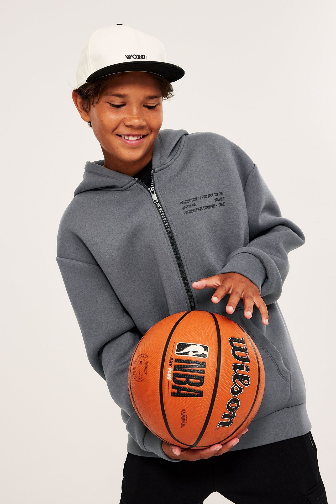 Dziecko w szarej bluzie scuba oversize z czarnym zamkiem, nadrukiem, białej czapce, trzymające piłkę.