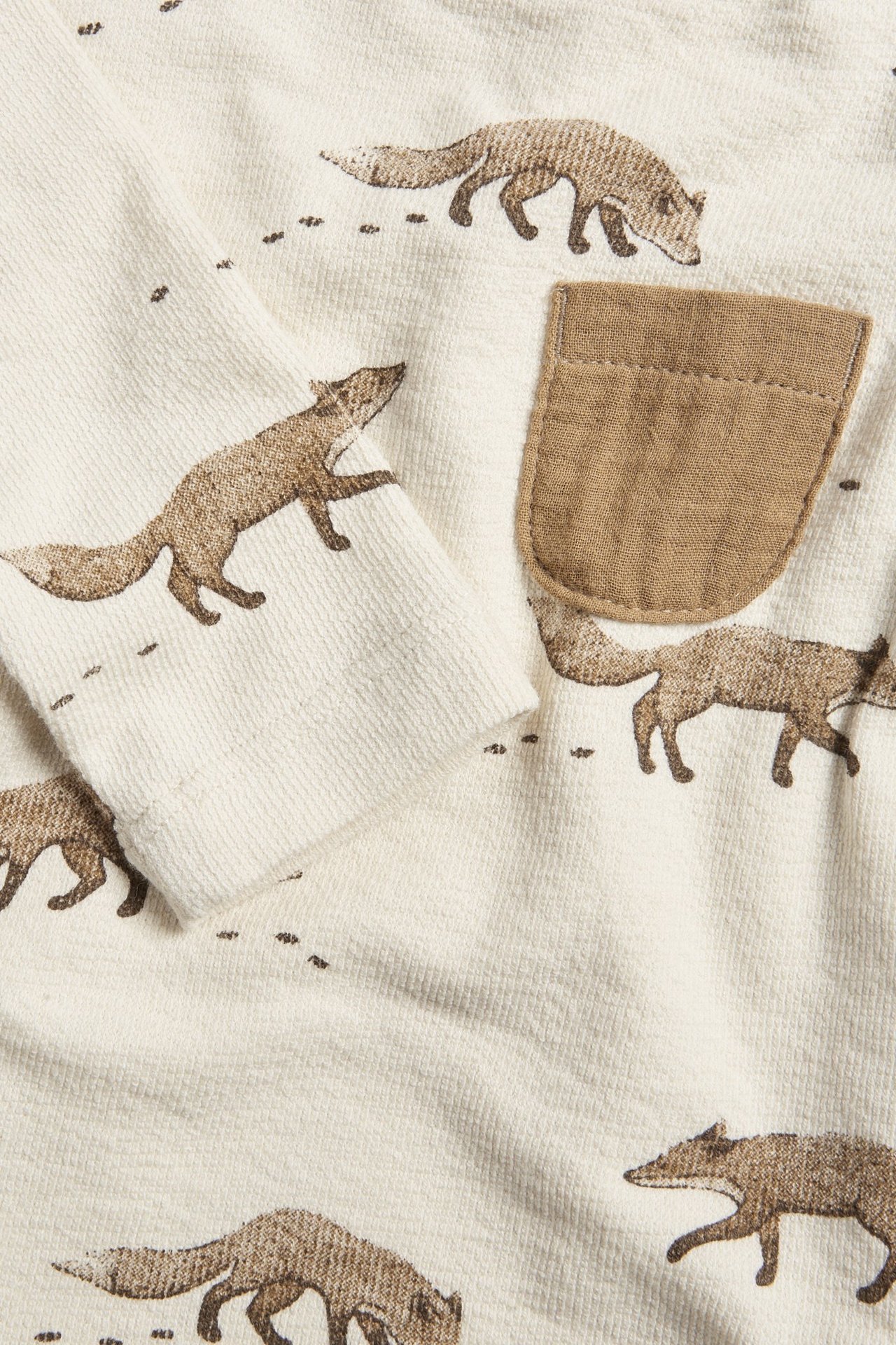 Nærbilde av en lys beige langermet babybody med brunt revemønster og en liten brystlomme.