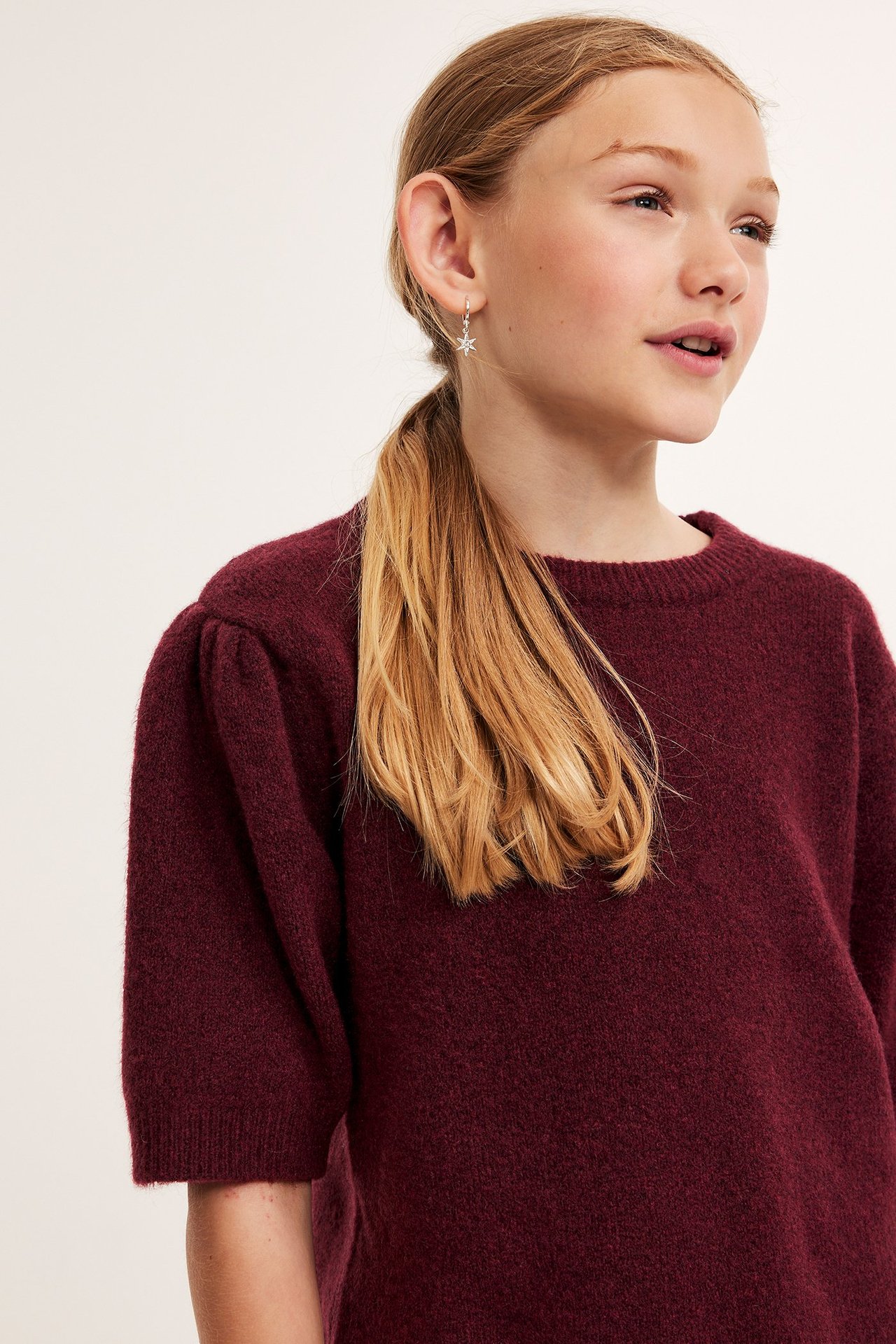 Zbliżenie, widok z boku dziecka w bordowym melanżowym dzianinowym T-shircie z bufiastymi rękawami i kolczykami-gwiazdkami.