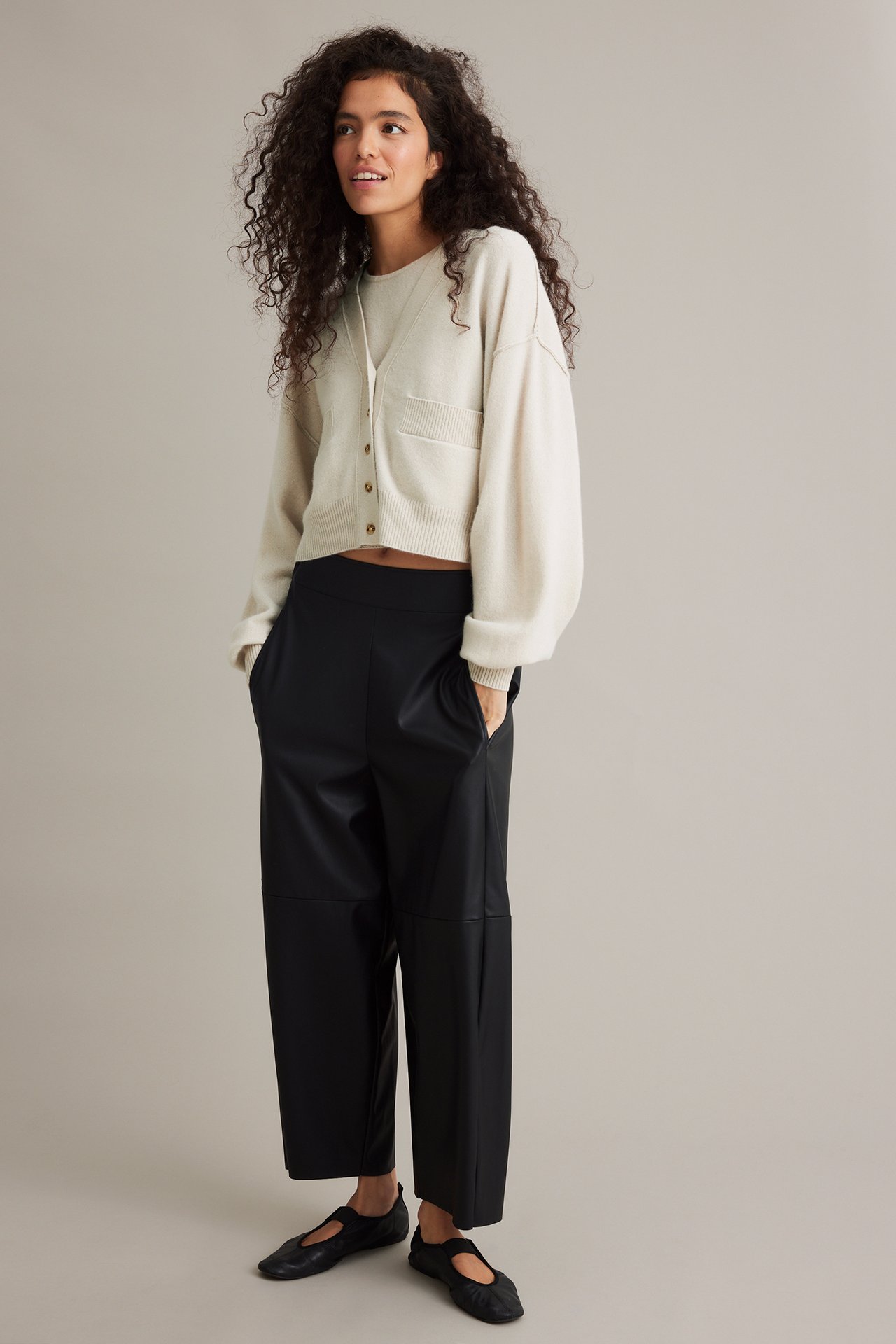 Front view: Kvinne har på seg beige kort cardigan, beige topp, sorte skinnimitasjonsbukser og sorte ballerinasko.