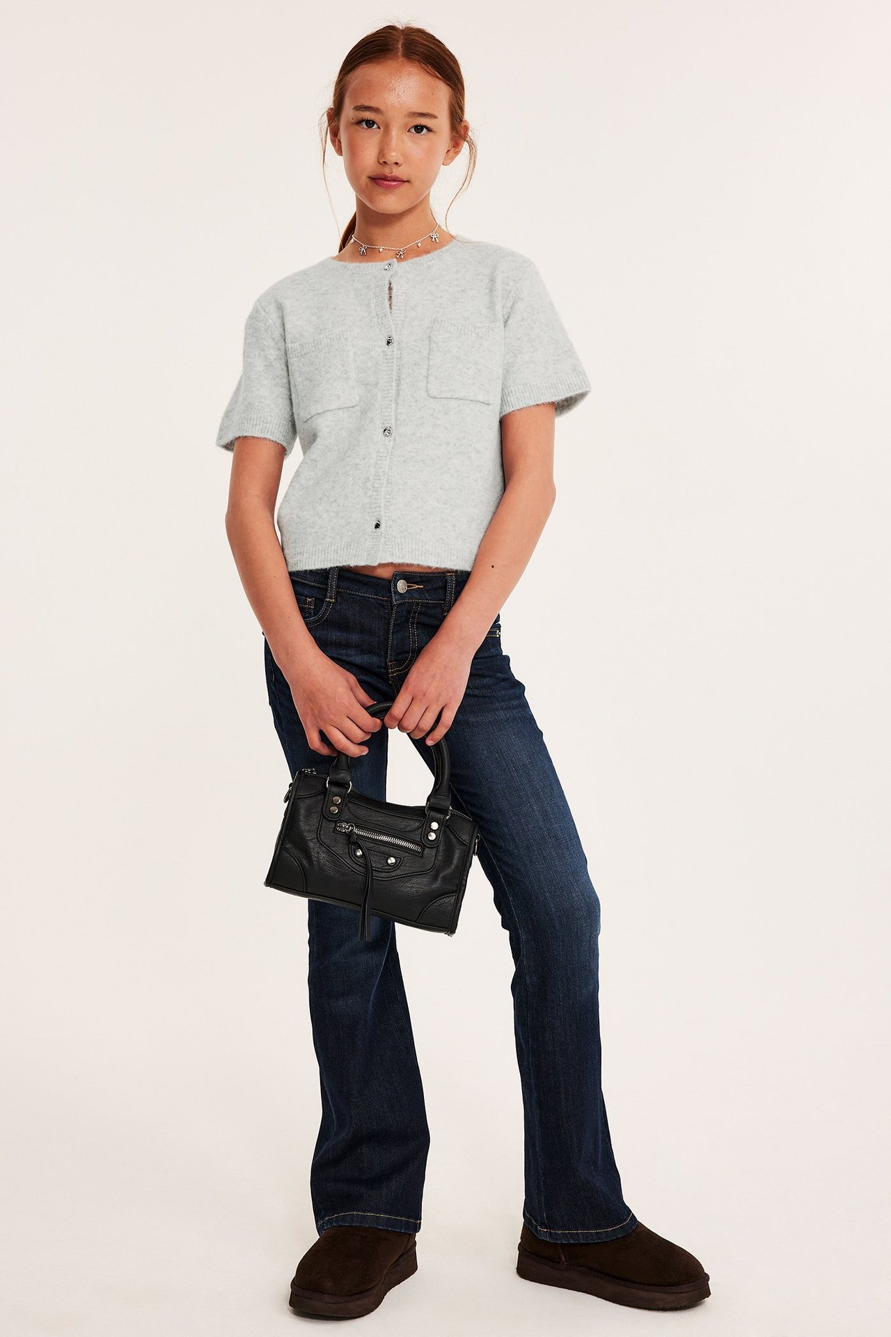 Widok z przodu: Dziecko w jasnoszarym melanżowym kardiganie, ciemnych dżinsach, trzyma czarną torebkę.