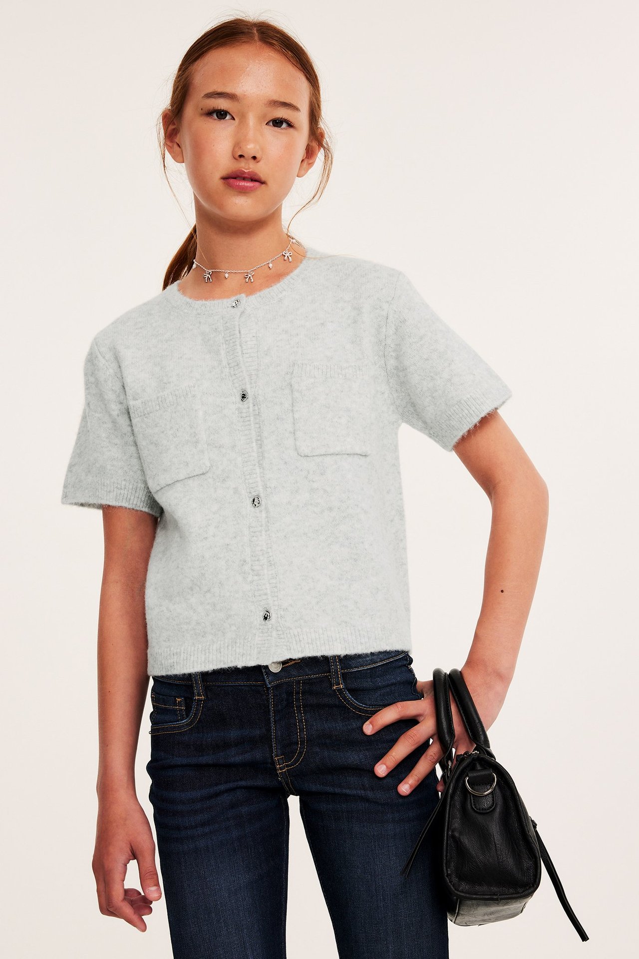 Widok z przodu: Dziecko w jasnoszarym melanżowym kardiganie, ciemnoniebieskich dżinsach, srebrnym naszyjniku, trzyma czarną torebkę.