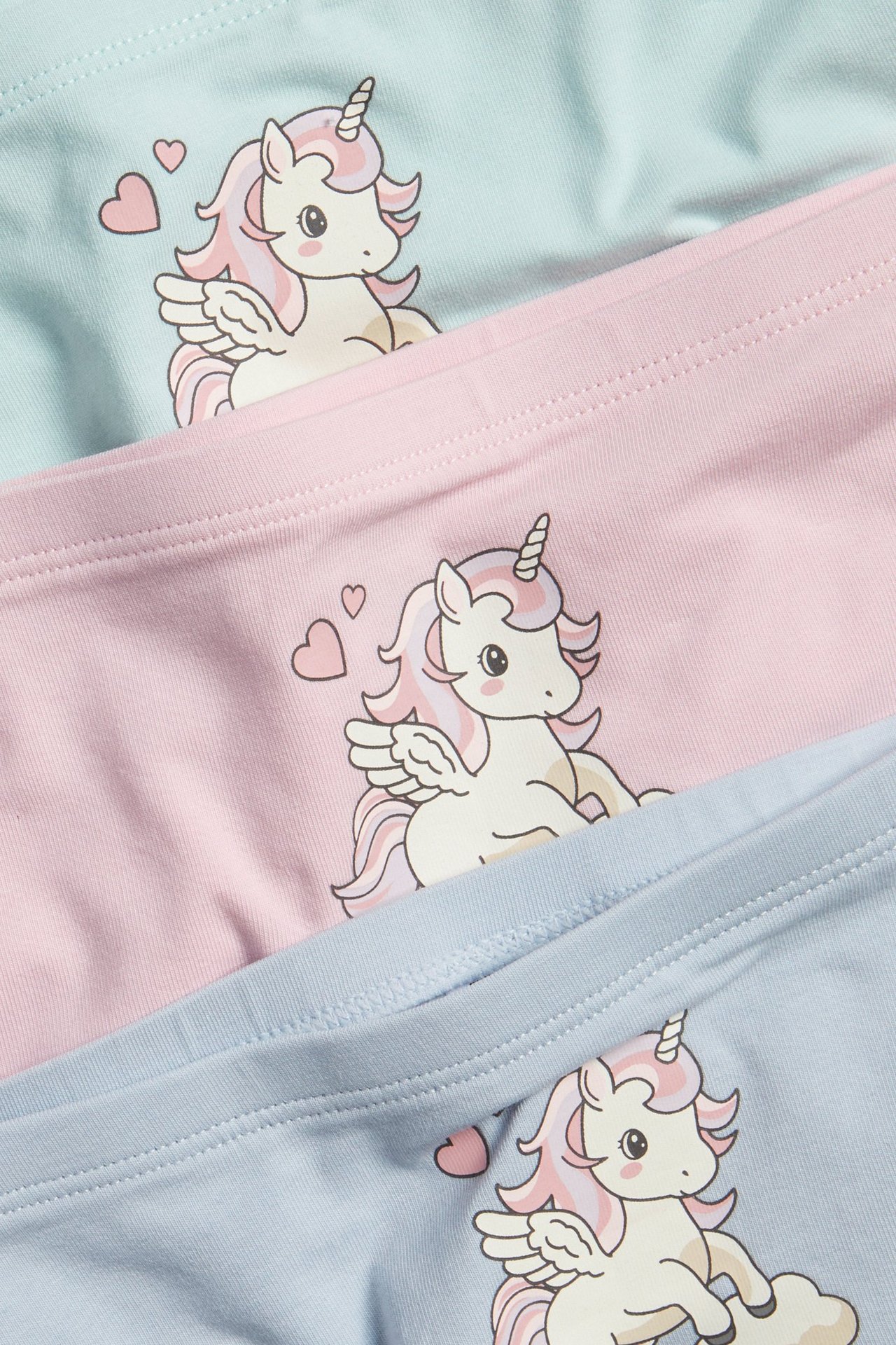 Zbliżenie trzech par dziecięcych bokserek w jasnoniebieskim, różowym i miętowym kolorze z jednorożcami.