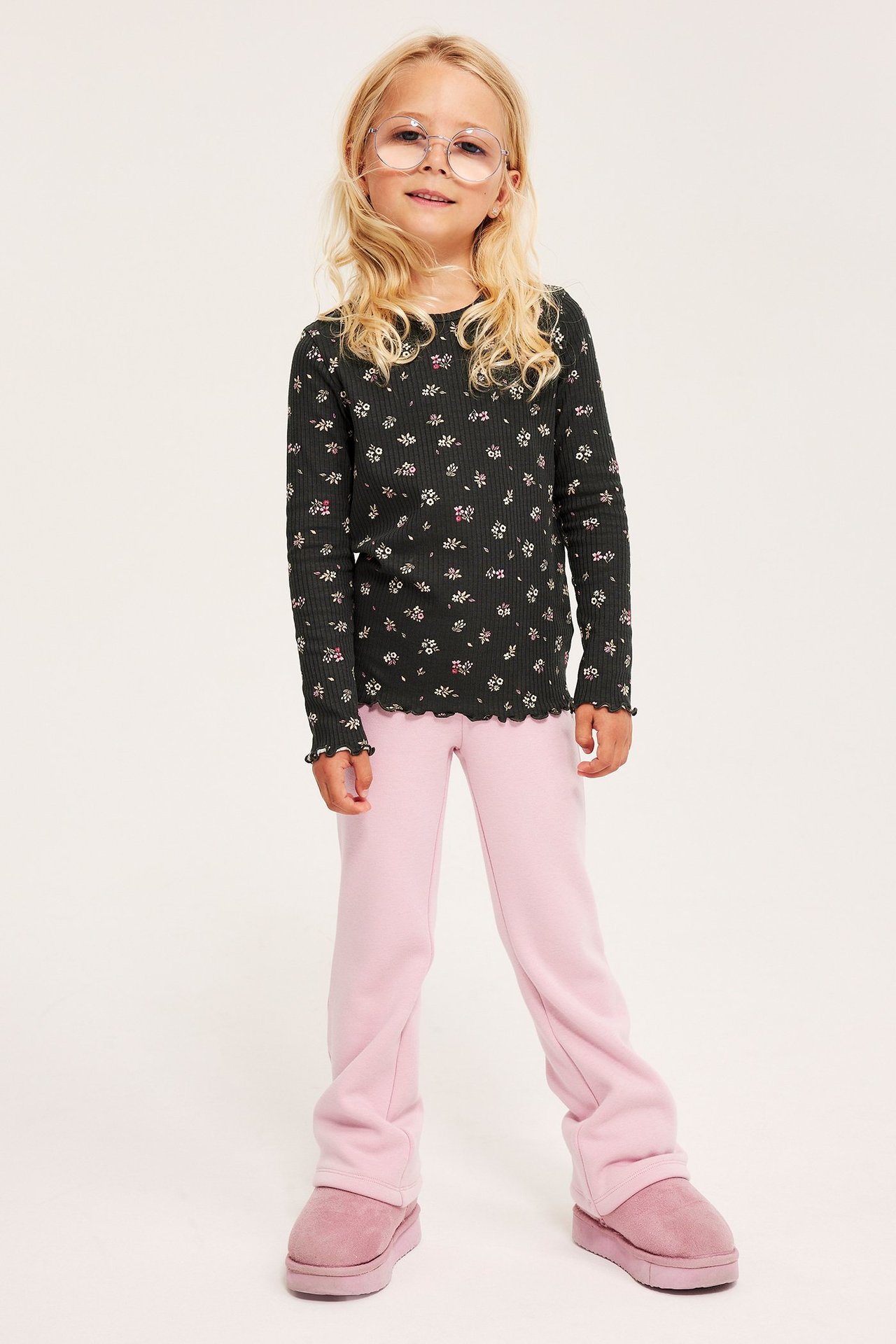 Forfra: barn i klare briller, svart ribbet blomstret langermet topp, rosa bukser og rosa støvler.