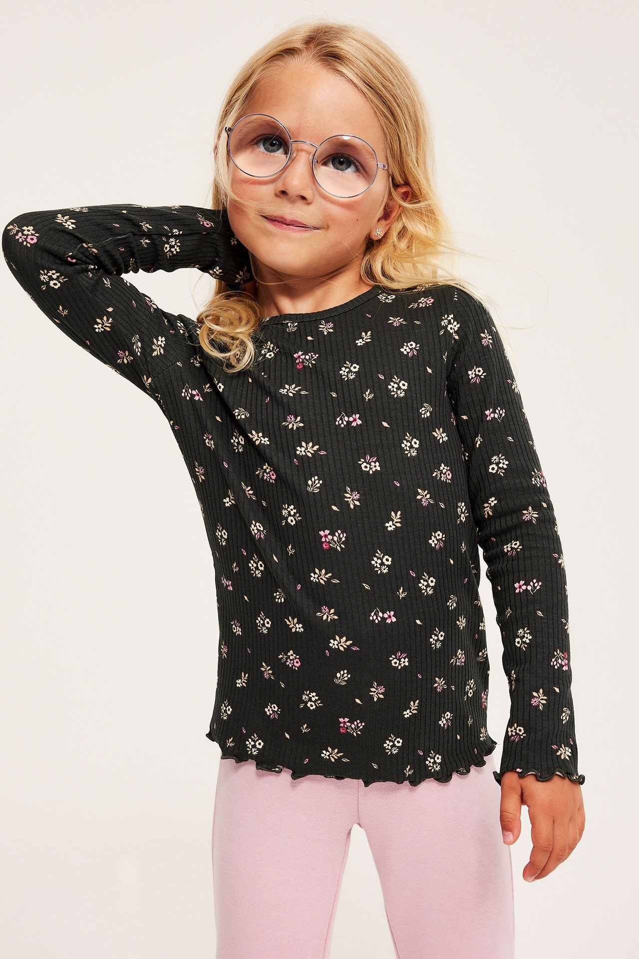 Forfra: Barn i svart ribbet langermet topp med blomstermønster, volangkanter, rosa leggings og briller.