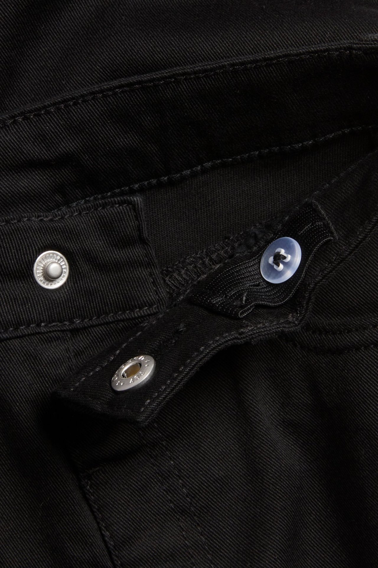 Lähikuva lasten mustien bootcut-farkkujen vyötäröstä, jossa painonappi ja säädettävä kuminauha napilla.