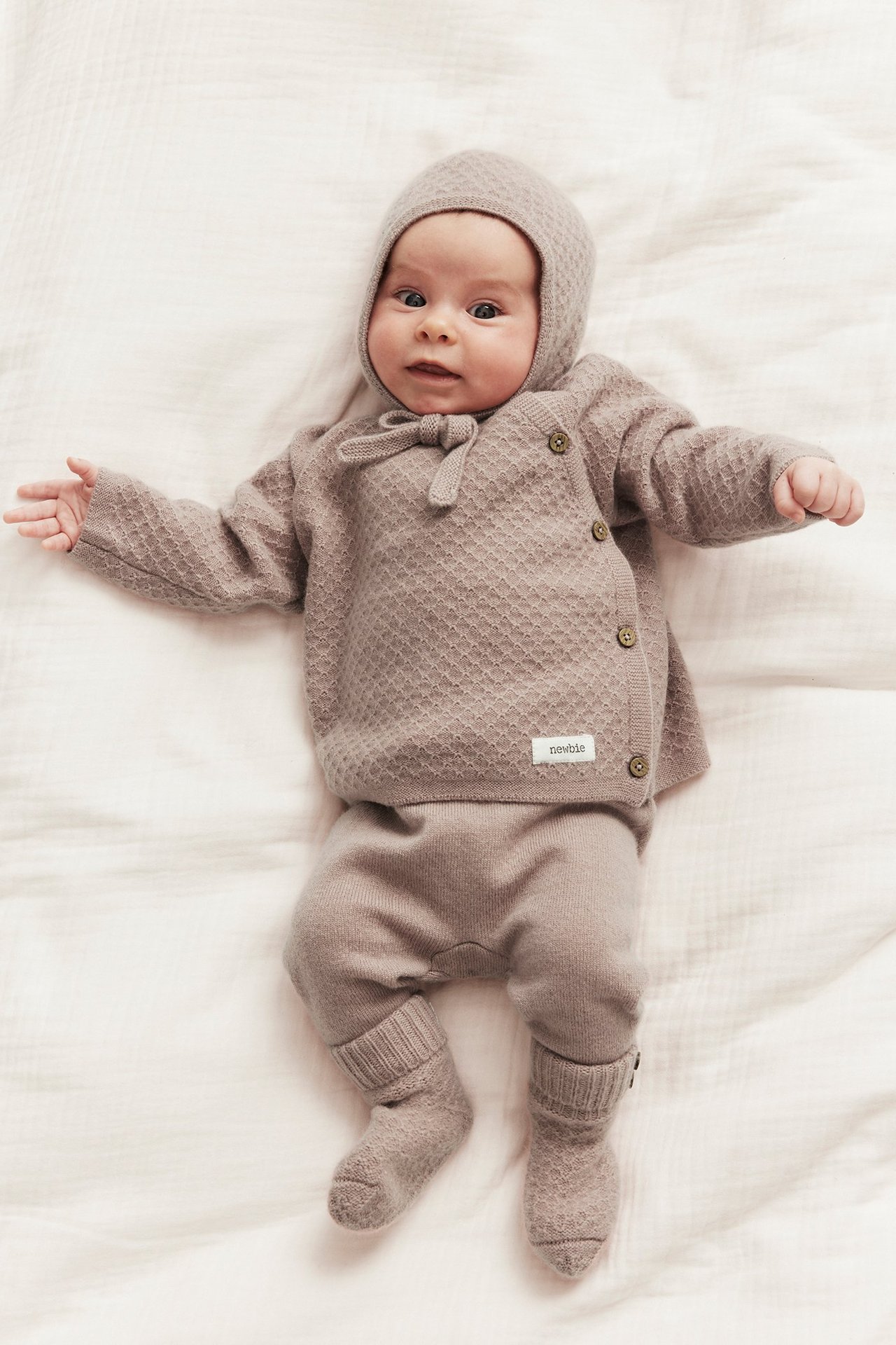 Forfra: baby i en lys rosa strukturstrikket ull-kasjmir cardigan med sideknapper, matchende lue, bukse og sokker.