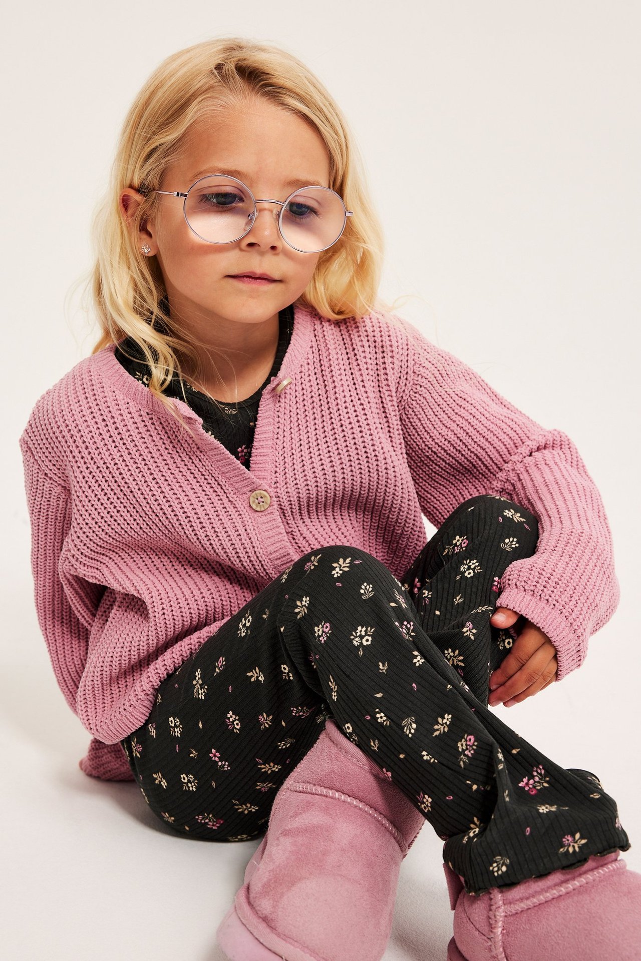 Dziecko z przodu w czarnych prążkowanych legginsach flare w kwiaty, różowym swetrze rozpinanym i różowych butach.