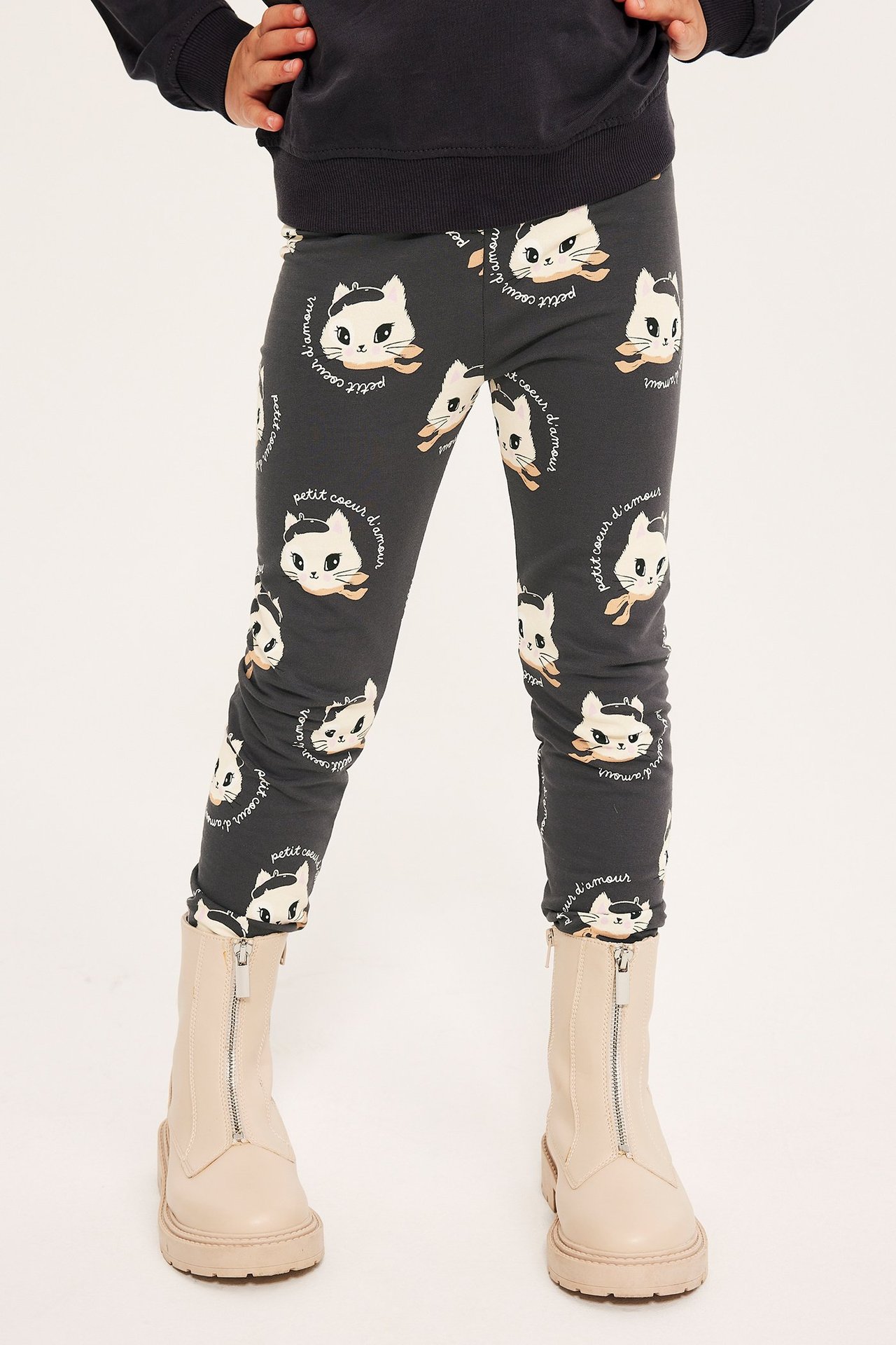 Forfra: et barn i mørkegrå mønstrede leggings med kattemotiv, en mørkegrå topp og lys beige støvler.