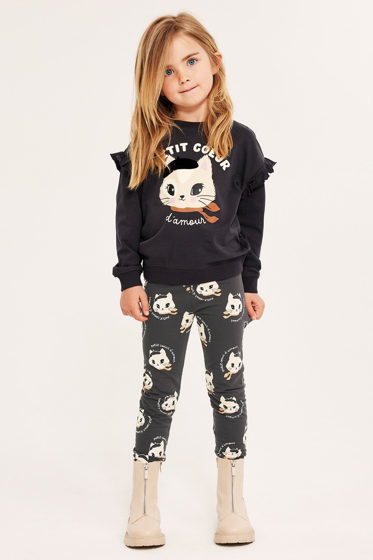 Widok z przodu dziecka w ciemnoszarych legginsach w koty, ciemnoszarej bluzie z kotem i beżowych botkach.