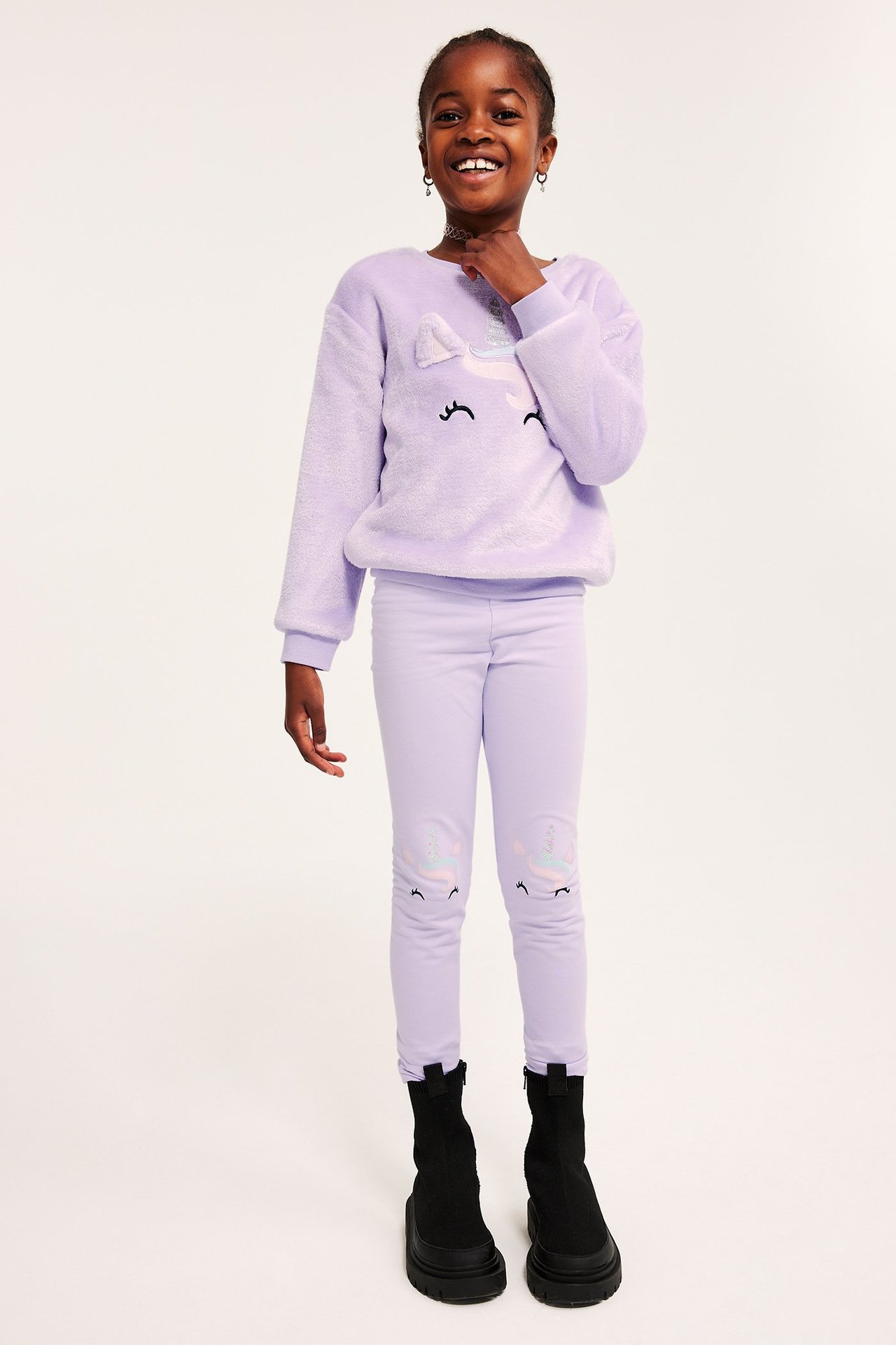 Frontbilde av et barn i lys lilla leggings med enhjørning-applikasjoner, en fluffy lilla genser og svarte støvler.