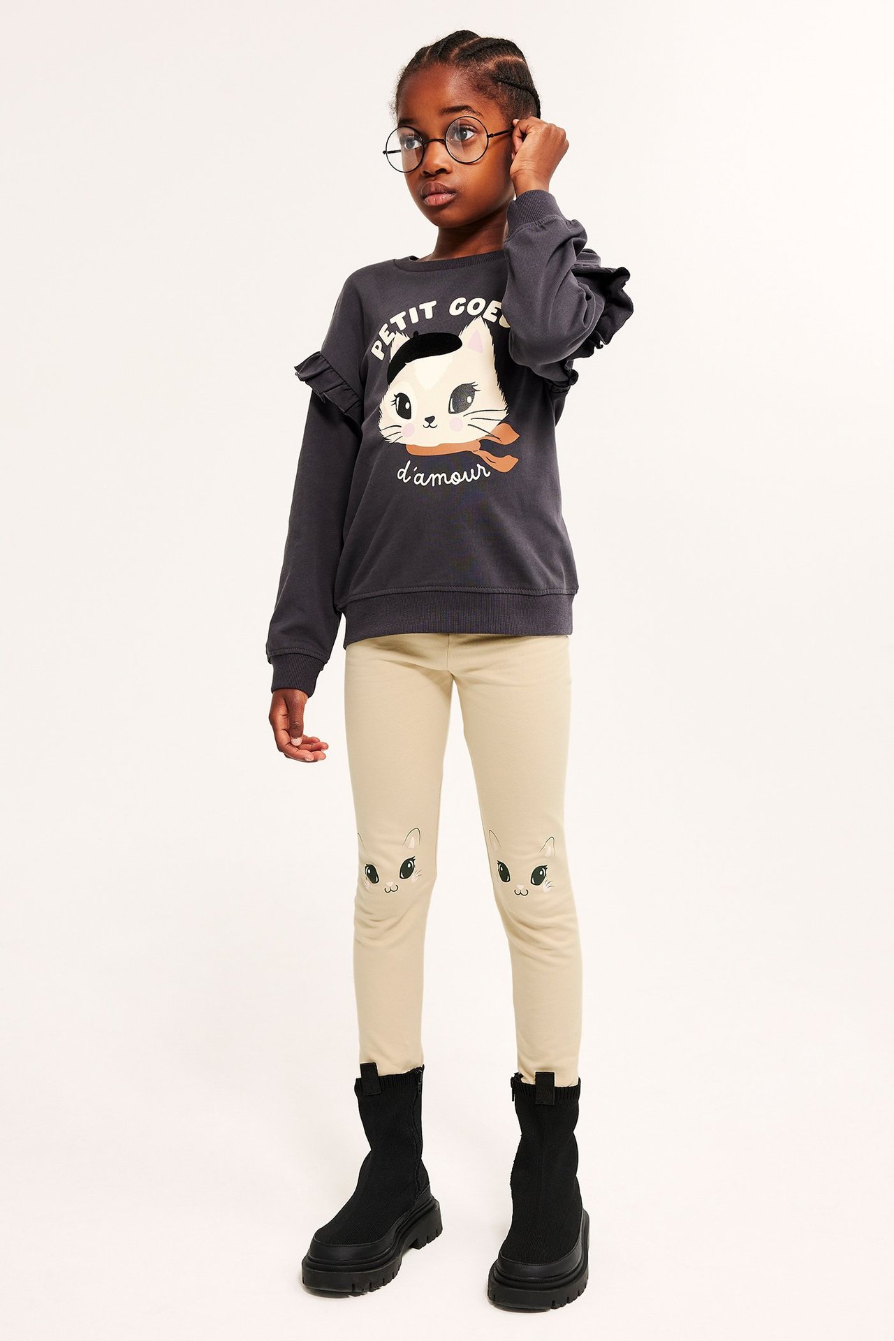 Edestä kuvattu lapsi beigessä leggingseissä kissakuvioilla, tummanharmaassa collegepaidassa ja mustissa saappaissa.