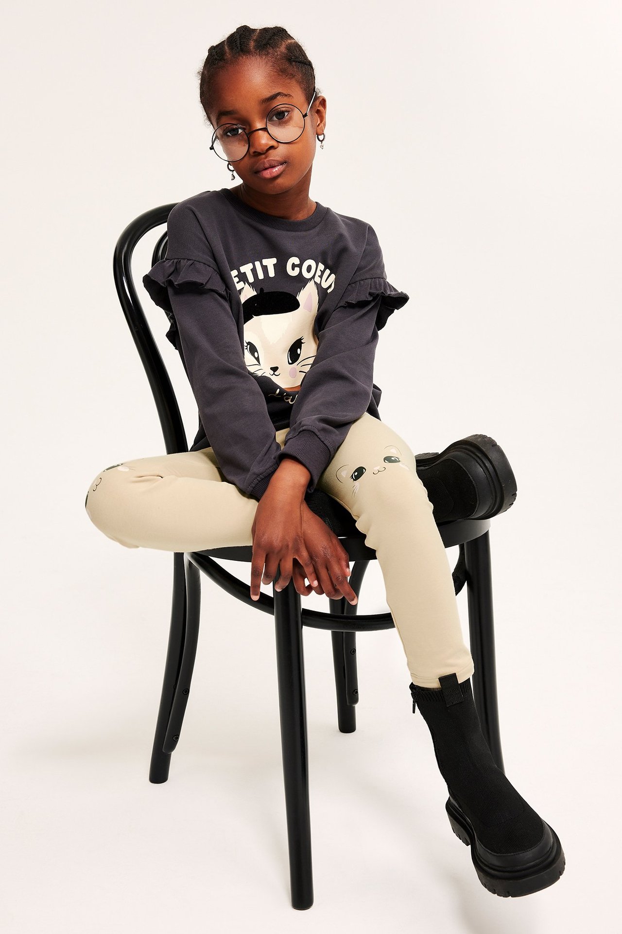 Widok z przodu dziecka w beżowych legginsach z aplikacjami kotów na kolanach, ciemnoszarym topie i czarnych butach.