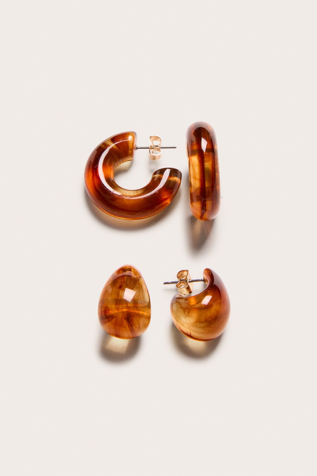 Lähikuva kahdesta parista ruskeita meripihkakuvioisia muovikorvakoruja: C-muotoiset renkaat ja pisaranmuotoiset tapit.