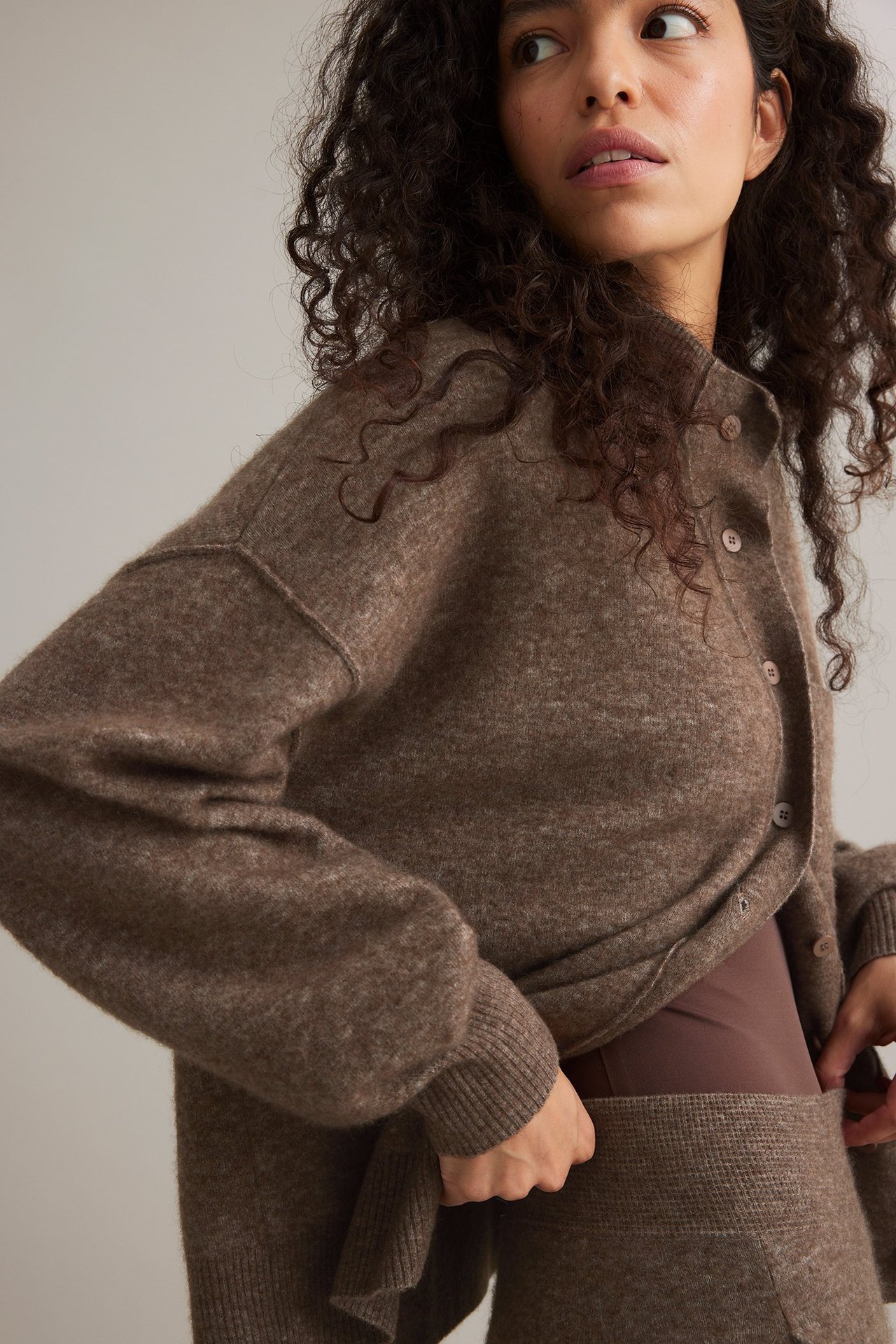Frontvy av en kvinna i öppen brunmelerad kofta i ullblandning över brun topp och matchande kjol.