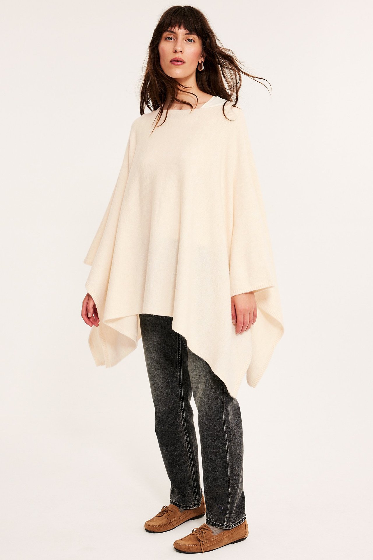 Frontbilde av en kvinne i offwhite strikket poncho over hvit topp, mørkegrå jeans og brune mokkasiner.
