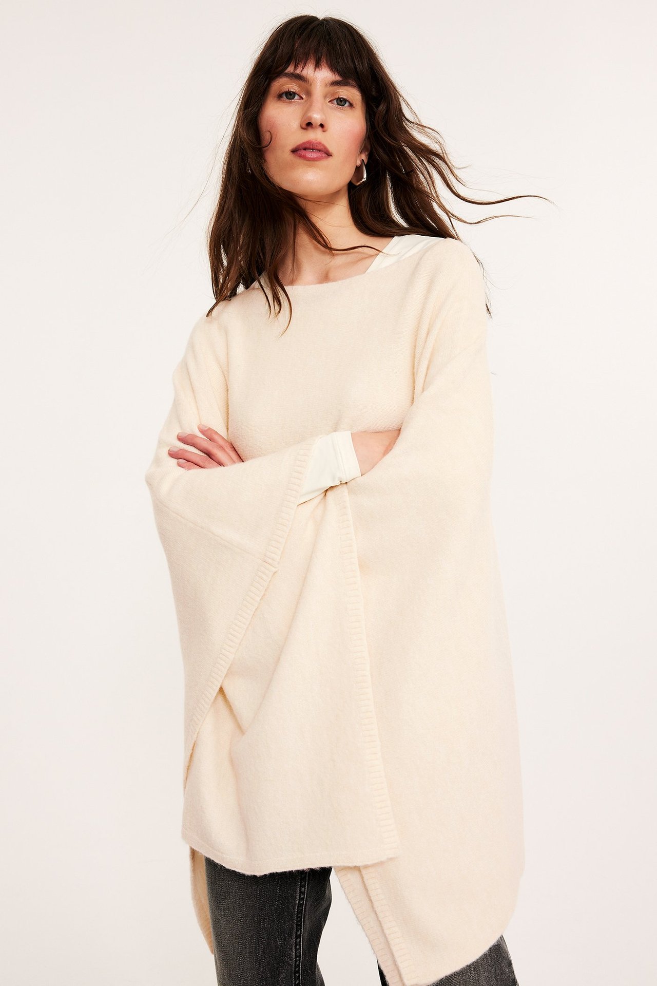 Frontbilde av en kvinne i en offwhite, løstsittende poncho over en lys topp og mørke jeans.