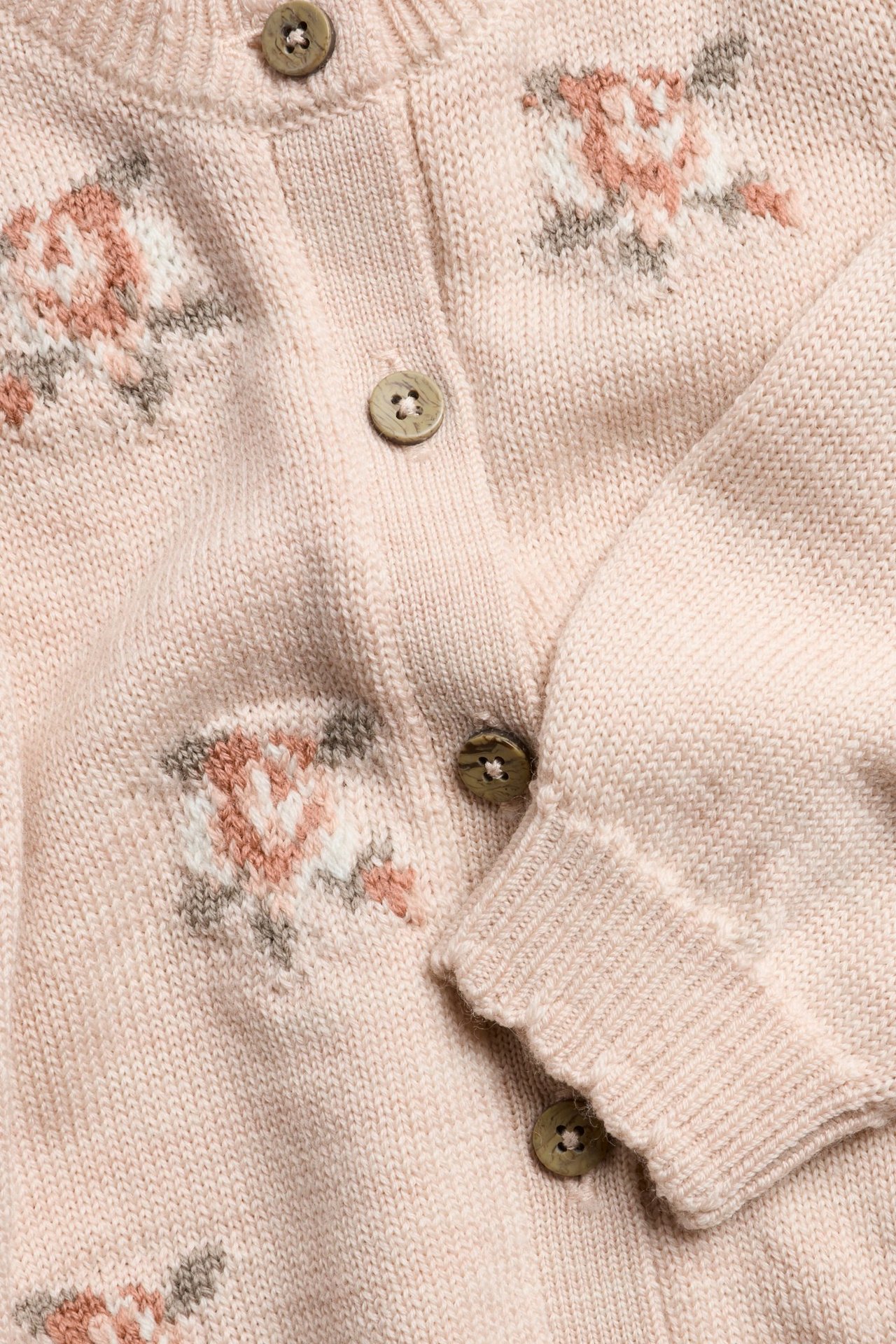 Närbild: rosa stickad ullkofta med jacquardrosor, bruna knappar, ribbade uddkanter.
