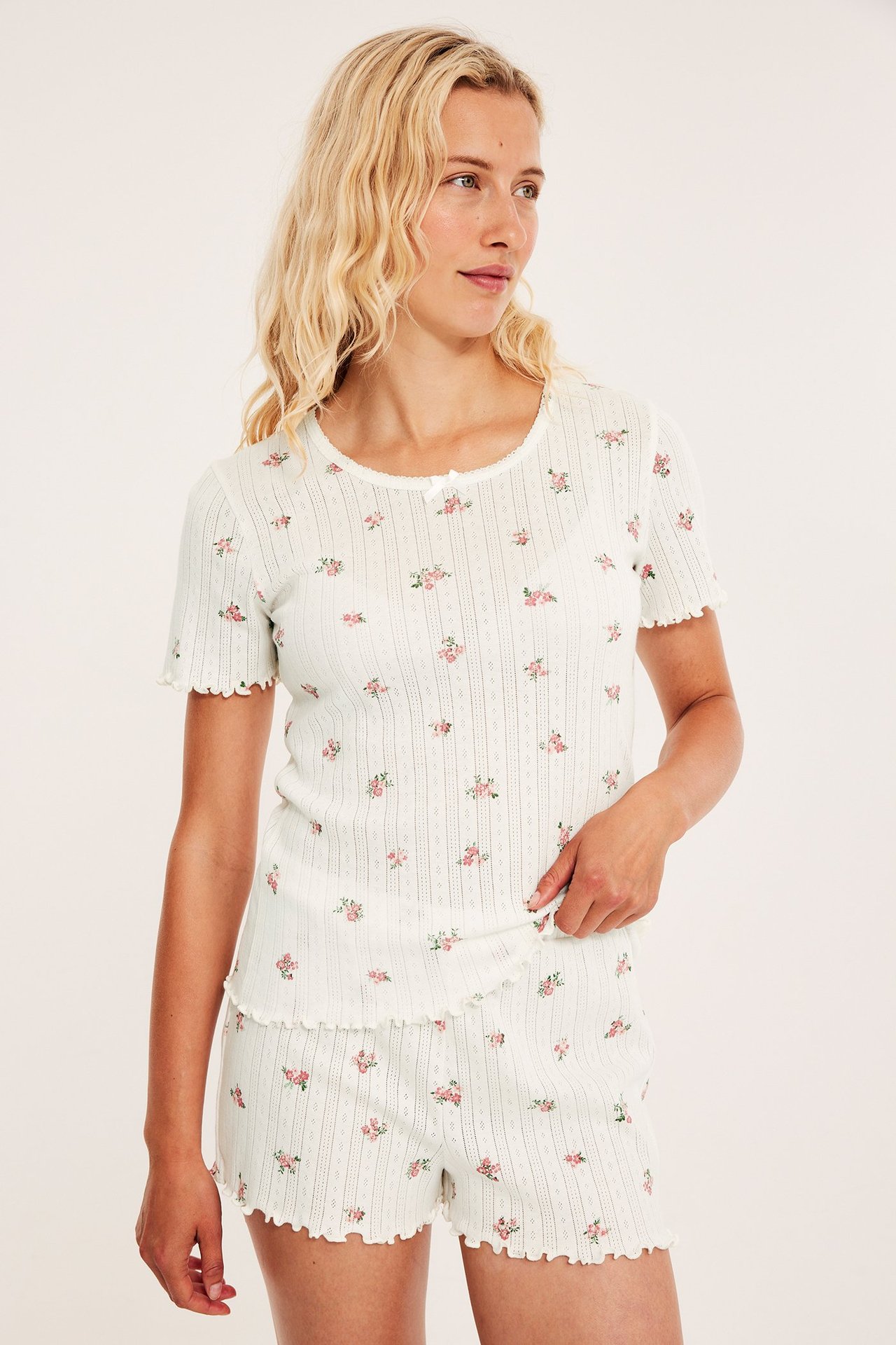 Edestä: nainen luonnonvalkoisessa pointelle-puuvillapyjamassa, jossa vaaleanpunainen kukkakuvio ja röyhelöreunat.