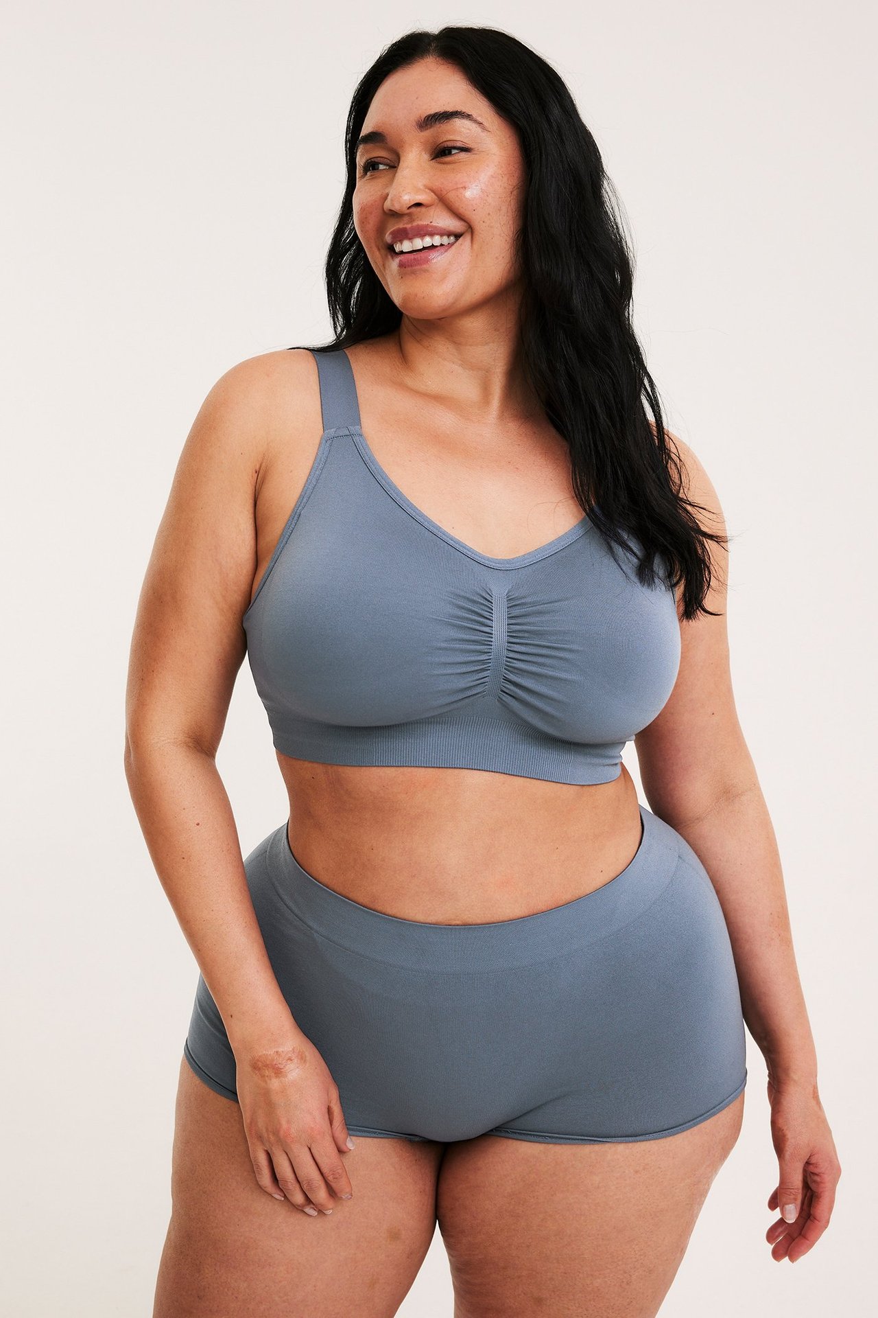 Hymyilevä nainen, edestä, tummansinisessä saumattomassa kaarituettomassa rintaliivissä rypytetyllä etuosalla ja yhteensopivissa shortseissa.