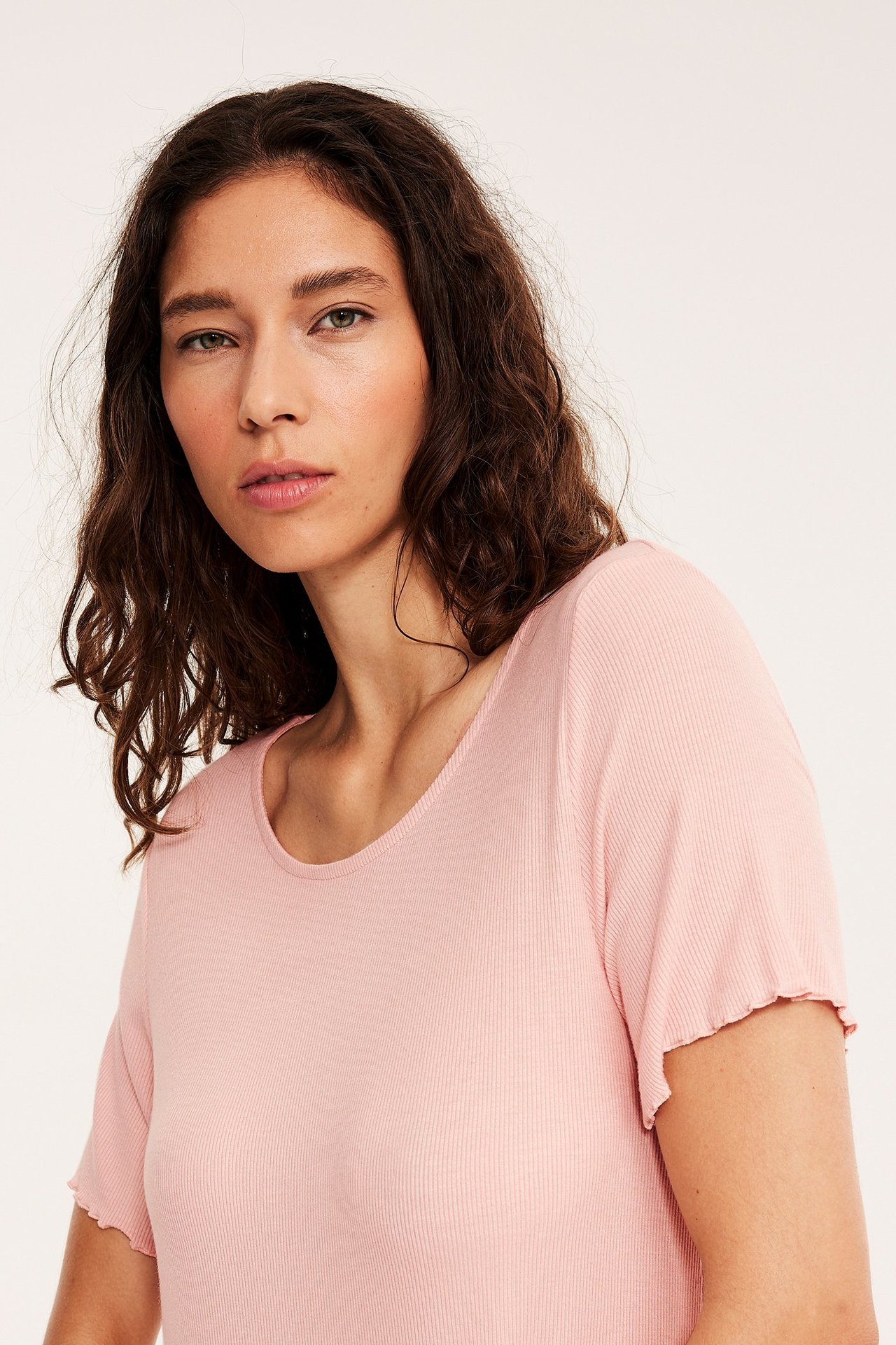 Lähikuva edestä naisesta, yllään vaaleanpunainen ribattu viskoosiyöpaita lyhyillä röyhelöhihoilla.