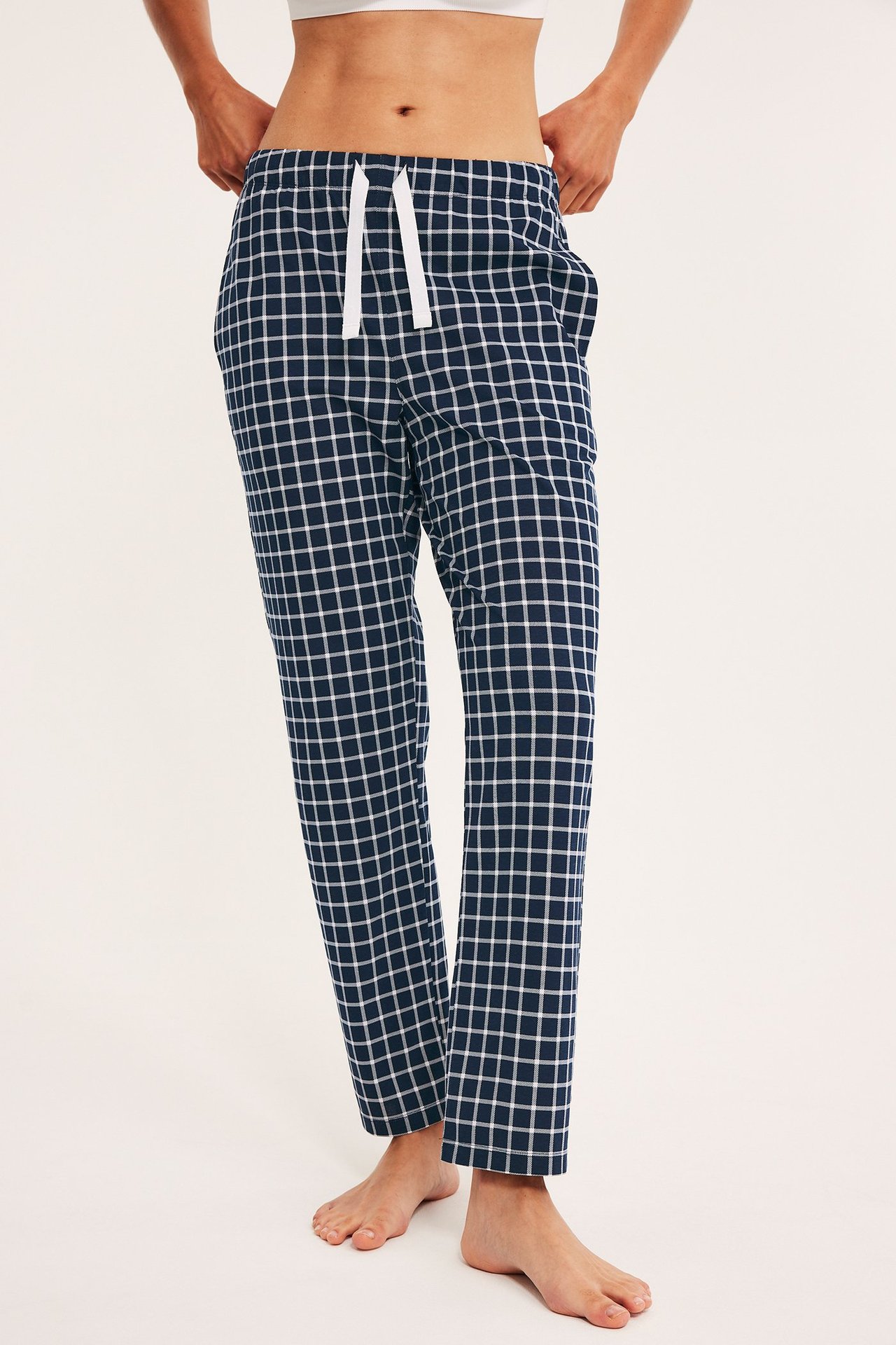 Edestä: nainen yllään tummansiniset ruudulliset puuvillapyjamahousut valkoisella kiristysnyörillä ja valkoinen toppi.