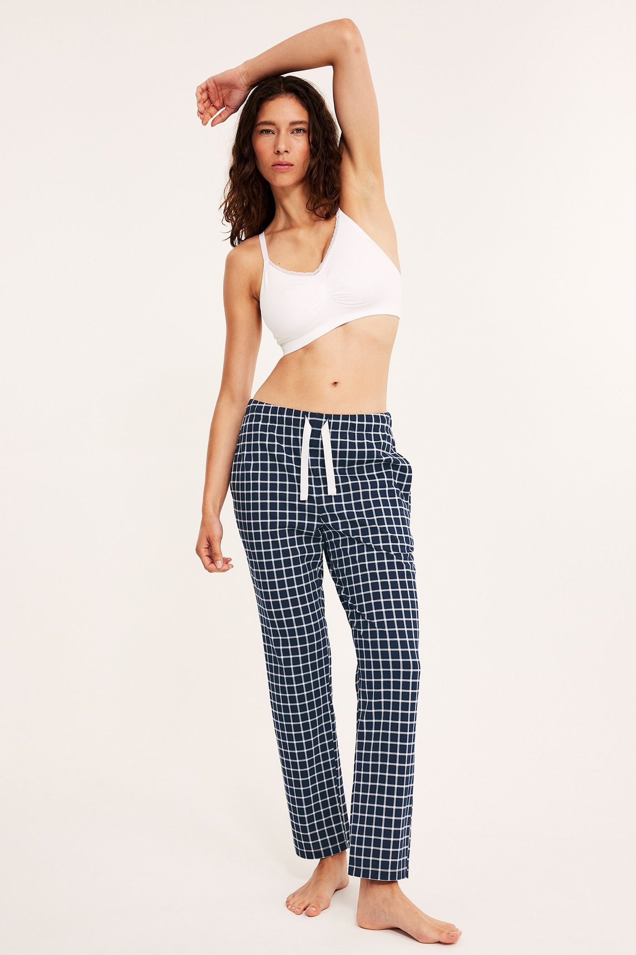 Widok z przodu: Kobieta w ciemnoniebieskich, kraciastych bawełnianych spodniach piżamowych i białym biustonoszu.