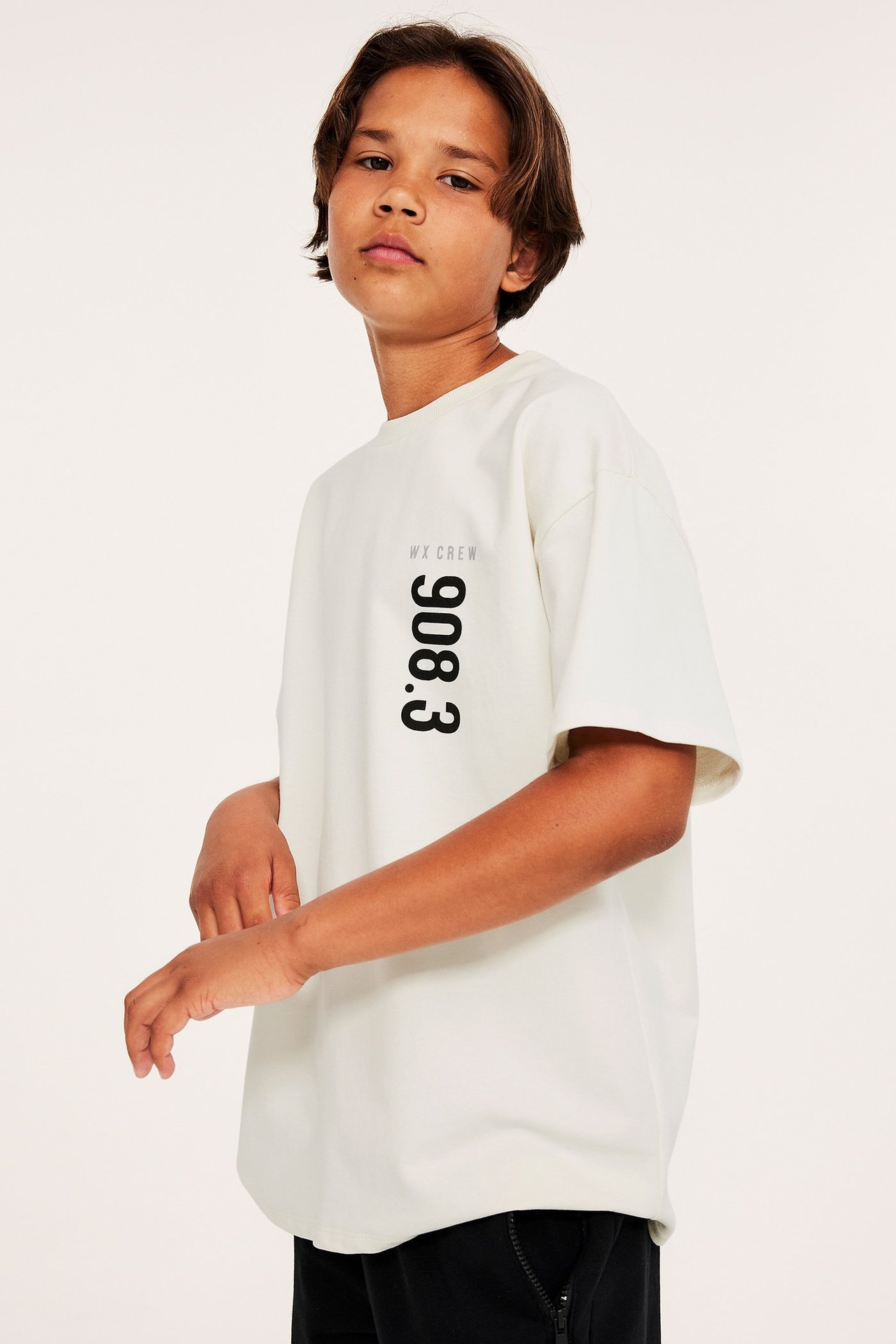 Frontvy av ett barn i en offwhite oversized bomulls-T-shirt med svart brösttryck och svarta byxor.