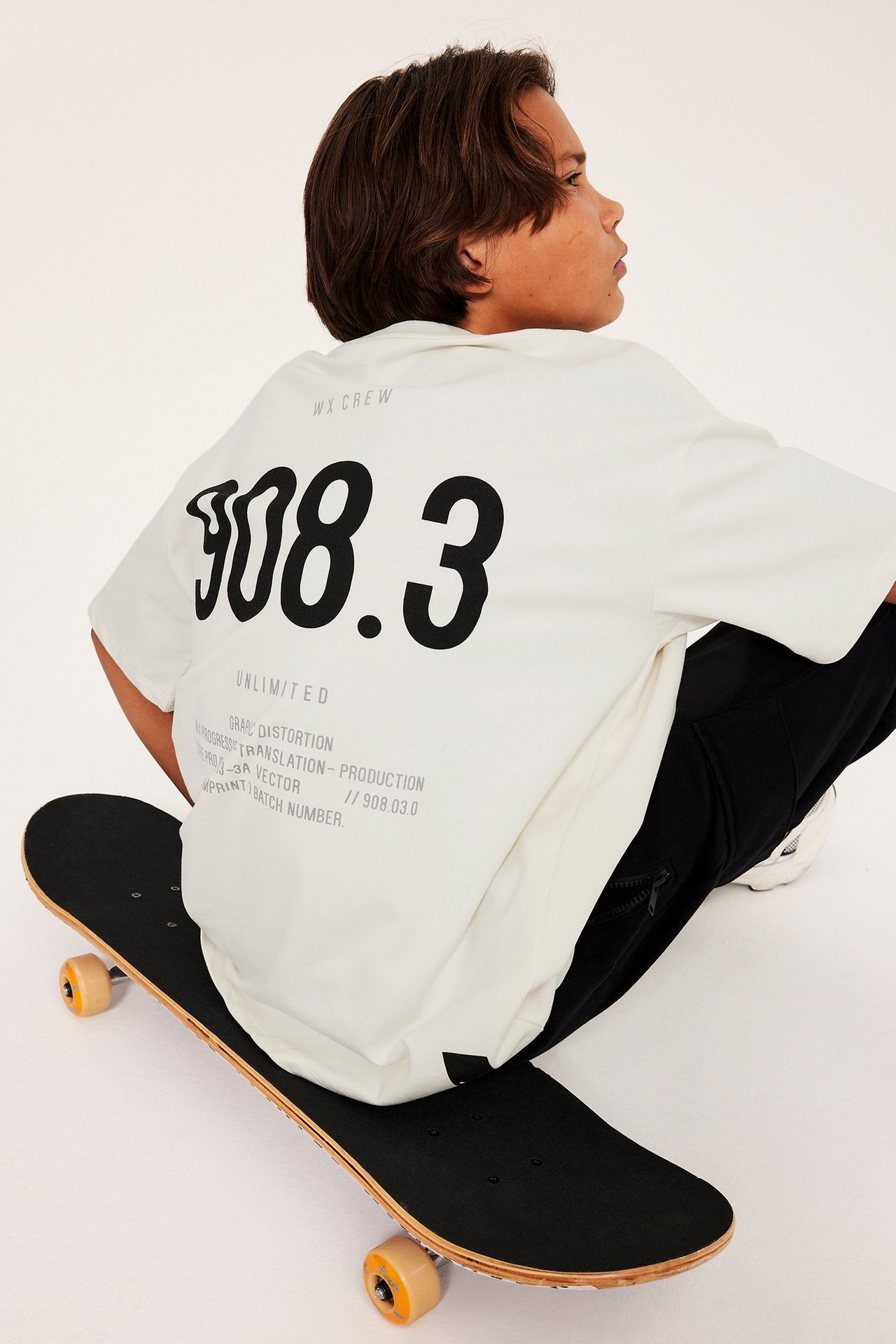 Bakifrån, ett barn i en offwhite oversized t-shirt med svart ryggtryck, sittande på en skateboard.