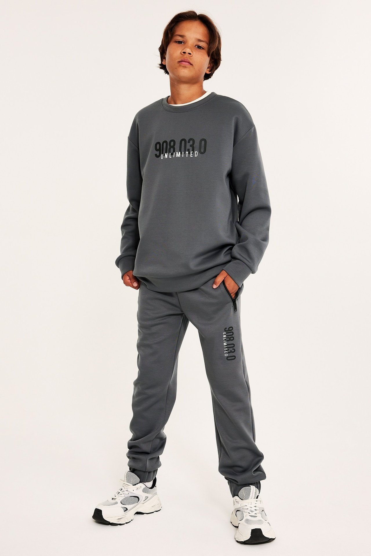Widok z przodu dziecka w ciemnoszarej bluzie z nadrukiem, pasujących dresach z zamkami i białych sneakersach.