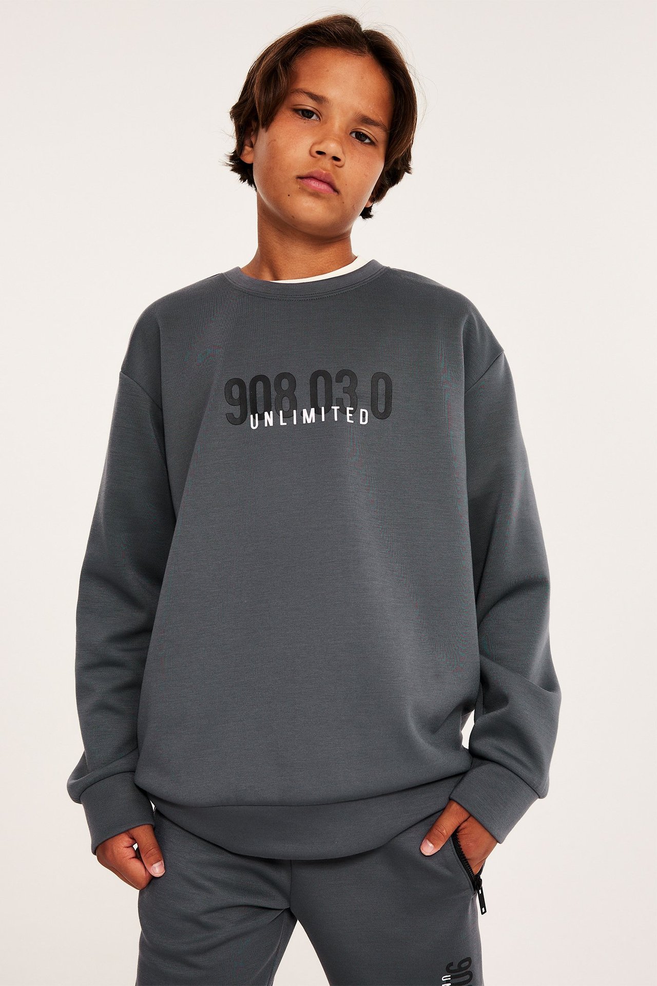 Dziecko, widok z przodu: Szara bluza dresowa z interlocku z nadrukiem '908.03.0 UNLIMITED', noszona z pasującymi szarymi spodniami.