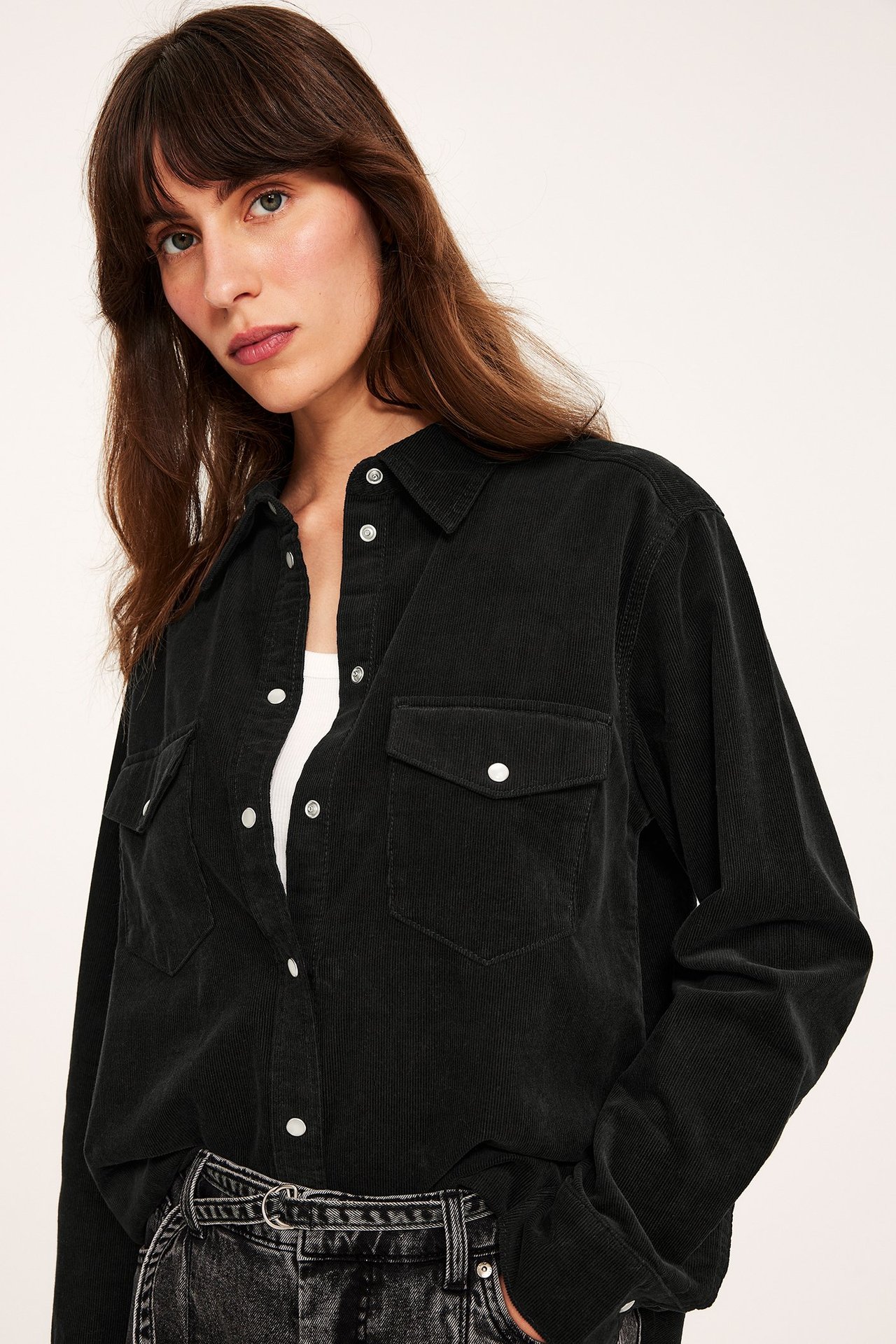 Widok z przodu kobiety w czarnej sztruksowej koszuli z zatrzaskami, na białym topie i ciemnoszarych dżinsach.