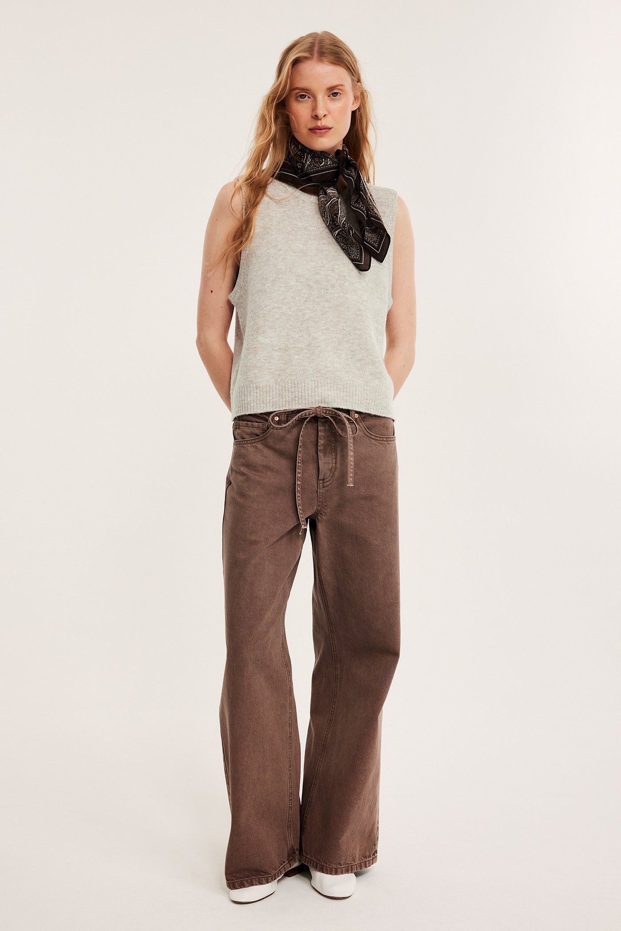 Framifrån: Kvinna i ljusbeige melerad stickad väst, bruna vida byxor, svart mönstrad scarf.