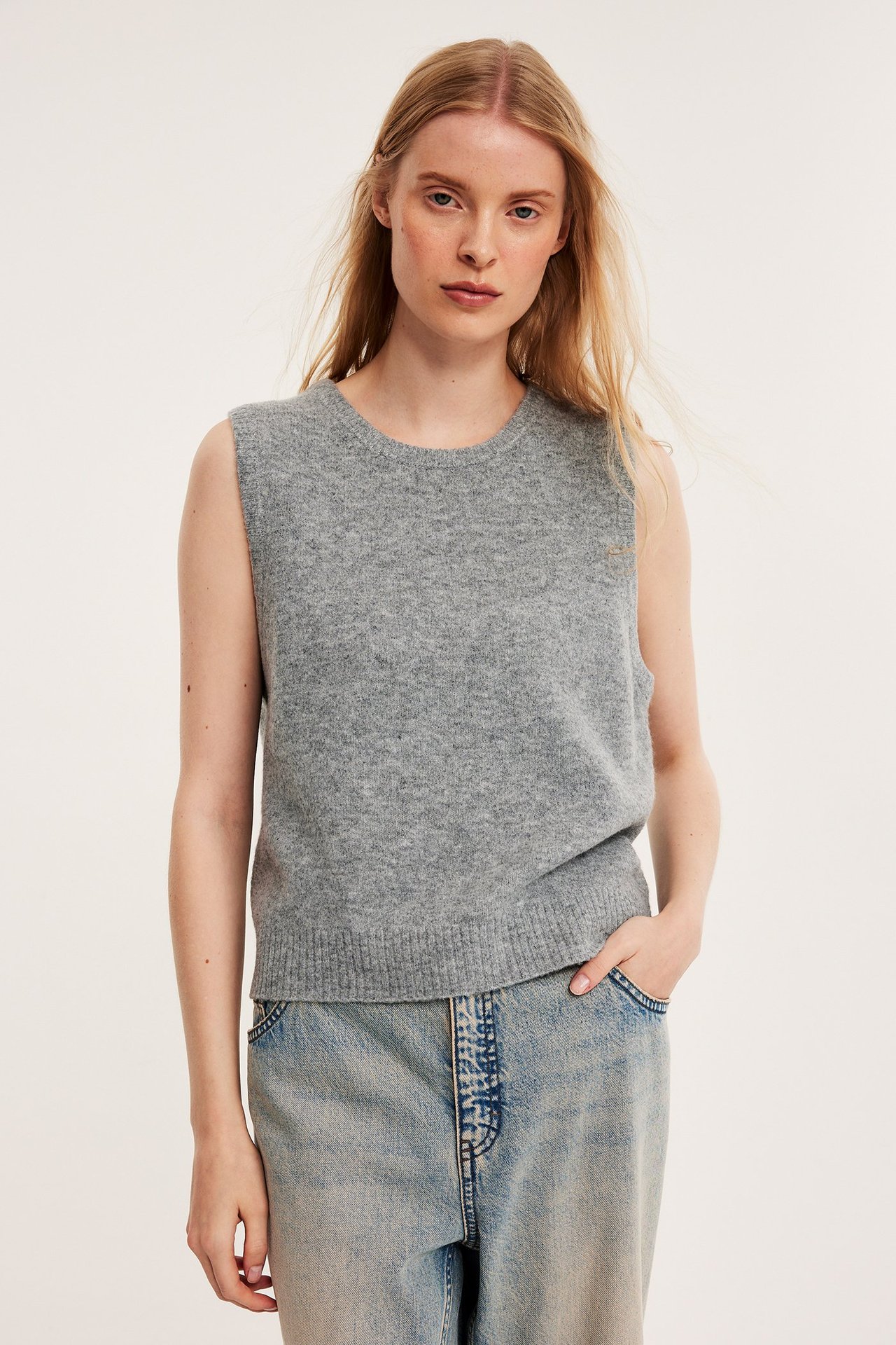 Framifrån: Kvinna i en ljusgrå melerad stickad väst och ljusblå jeans.