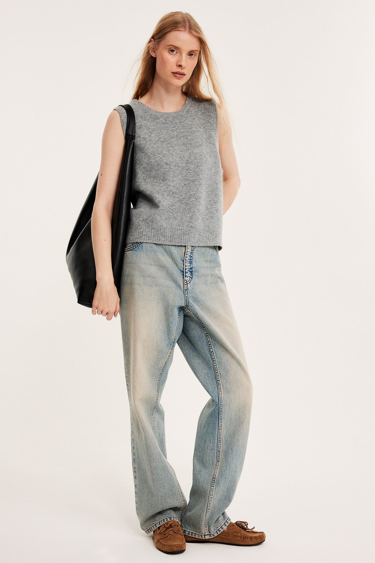 Framifrån: Kvinna i ljusgråmelerad stickad väst, ljusblå vida jeans, bruna mockasiner, håller svart väska.