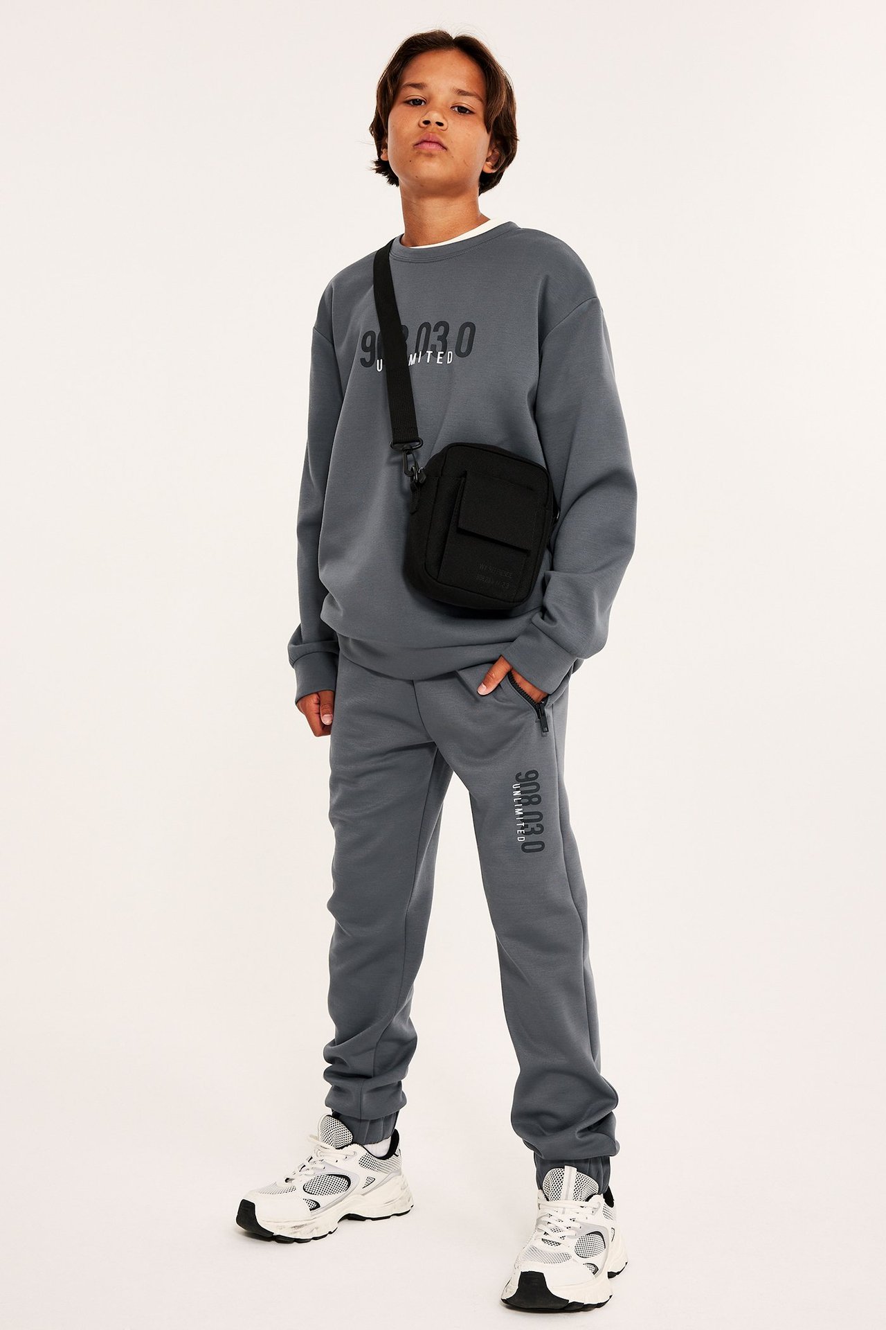 Dziecko: szare dresy z nadrukiem, szara bluza, czarna torba, białe sneakersy.