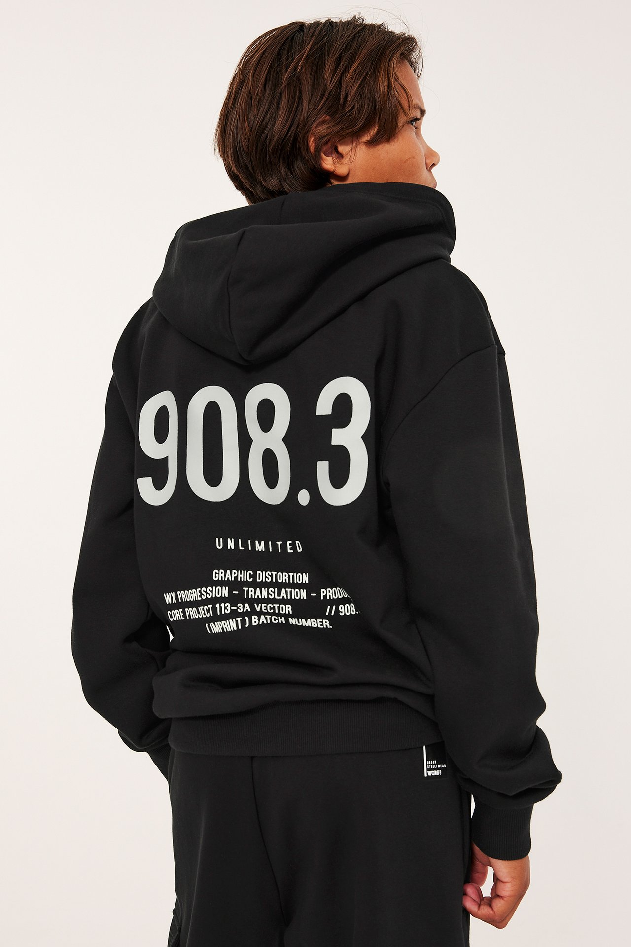 Bakifrån: ett barn i svart hoodie med vitt texttryck på ryggen och svarta mjukisbyxor.