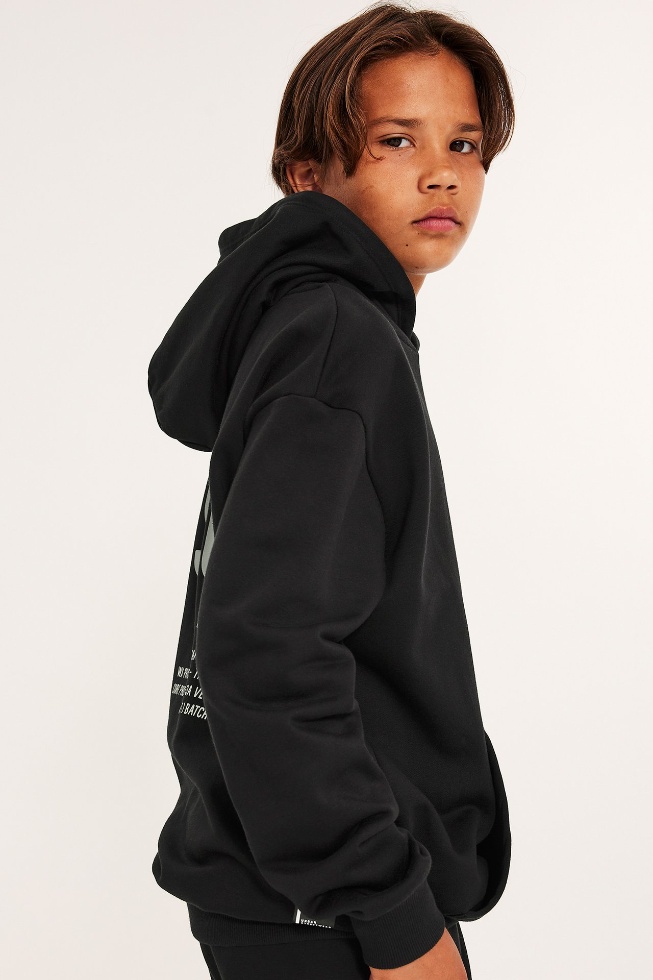 Sidovy av ett barn i en svart hoodie med vitt grafiskt tryck på ryggen, ribbade muddar.