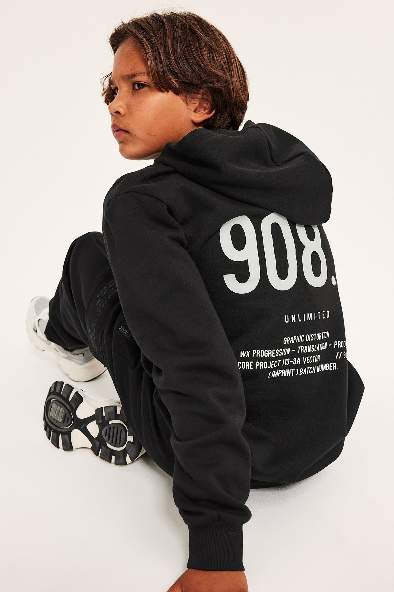 Widok z tyłu dziecka w czarnej bluzie z kapturem z białym nadrukiem, czarnych spodniach i białych sneakersach.